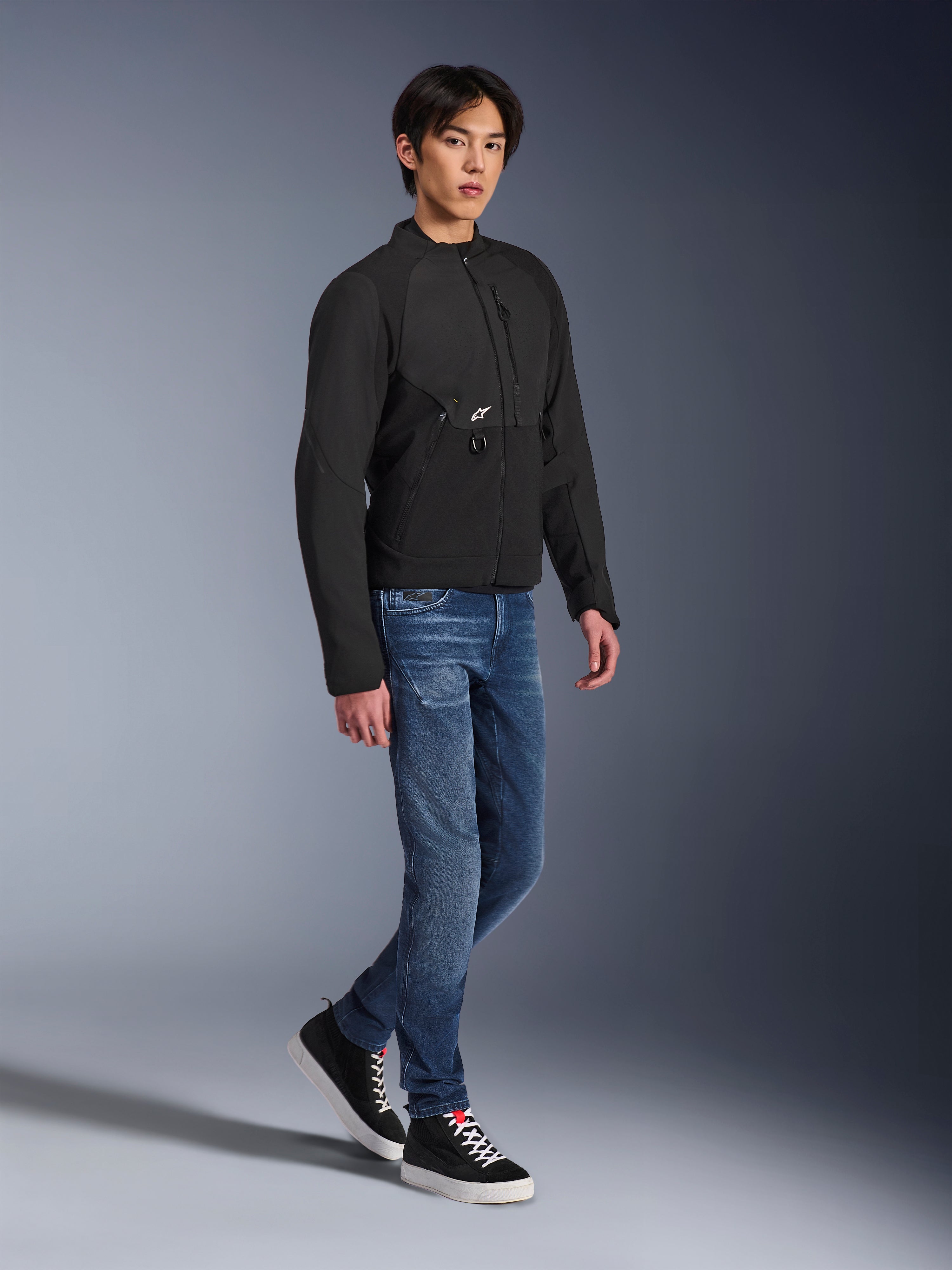 Alpinestars Provoke-jakke, urban motorcykeljakke, sort, båret af en mandlig model med blå jeans og sorte sneakers, teknisk tekstildesign med et lille Alpinestars-logo, fuldkrops livsstilsbillede til moderne bykørsel
