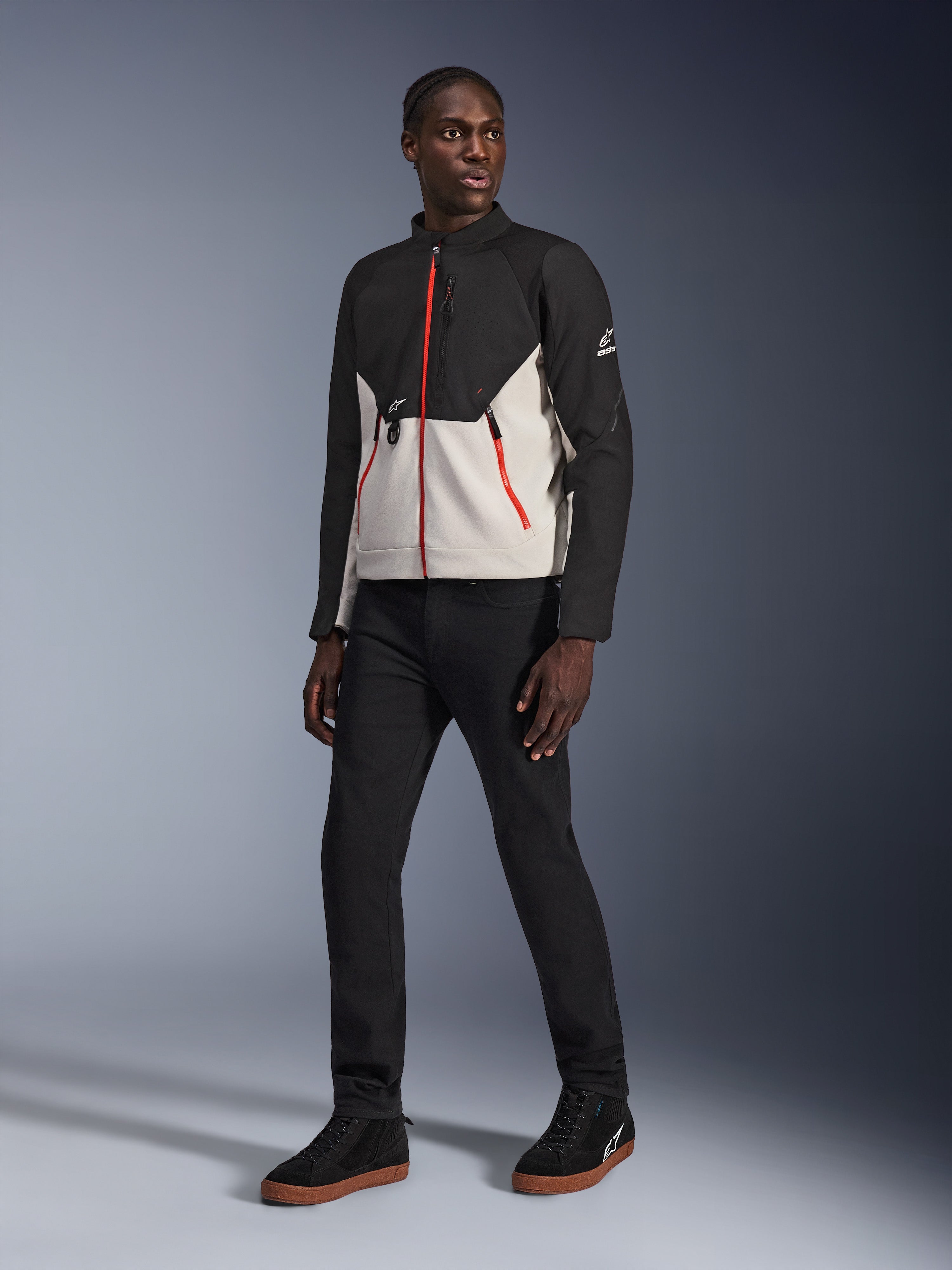 Model iført Alpinestars Provoke Jacket i Black Metal Gray, med karakteristiske røde lynlåse foran og på lommerne, kombineret med sorte bukser og sorte high-top sneakers med gummisåler, urbant moderne motorcykeludstyr til et street-ready look