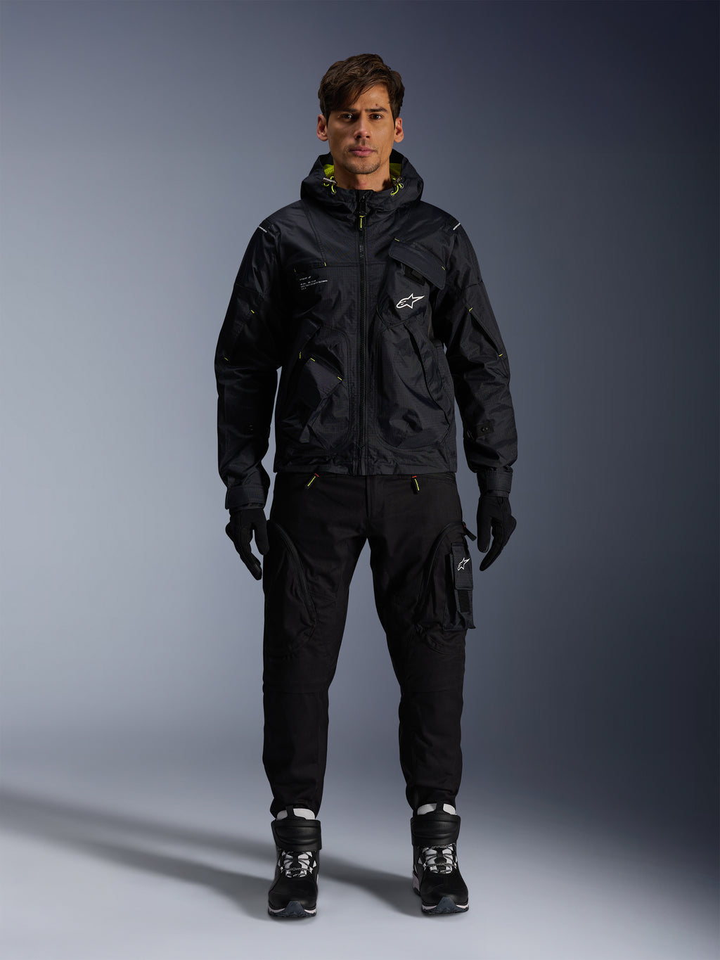 Mandlig model iført Alpinestars Mogress Air-jakke, sort tekstiljakke med hætte og lime punch-detaljer, sorte utility-bukser, sorte handsker og sort-hvide kørestøvler, urbant motorsport-inspireret streetwear-design