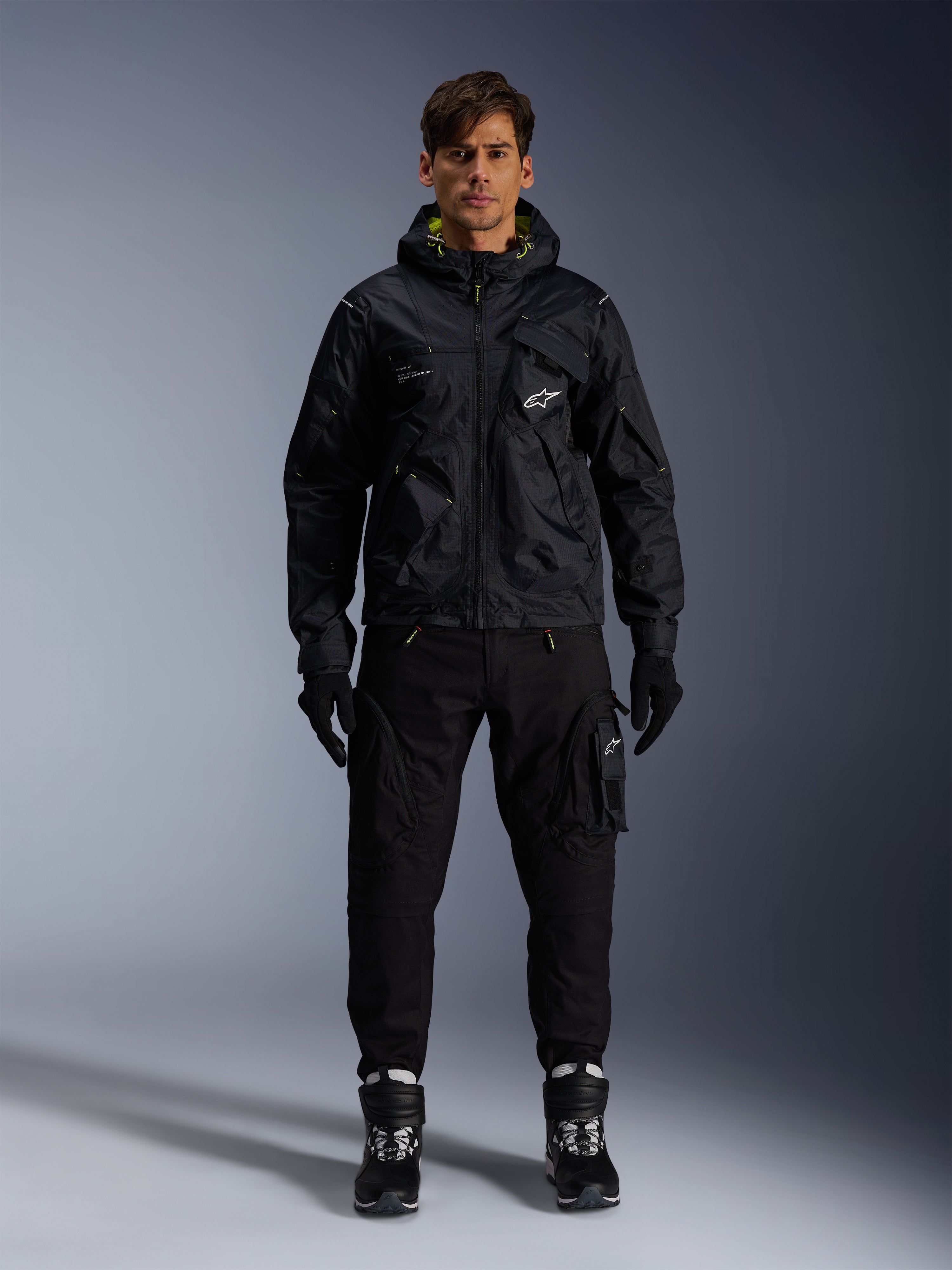 Mandlig model iført Alpinestars Mogress Air-jakke, sort tekstiljakke med hætte og lime punch-detaljer, sorte utility-bukser, sorte handsker og sort-hvide kørestøvler, urbant motorsport-inspireret streetwear-design