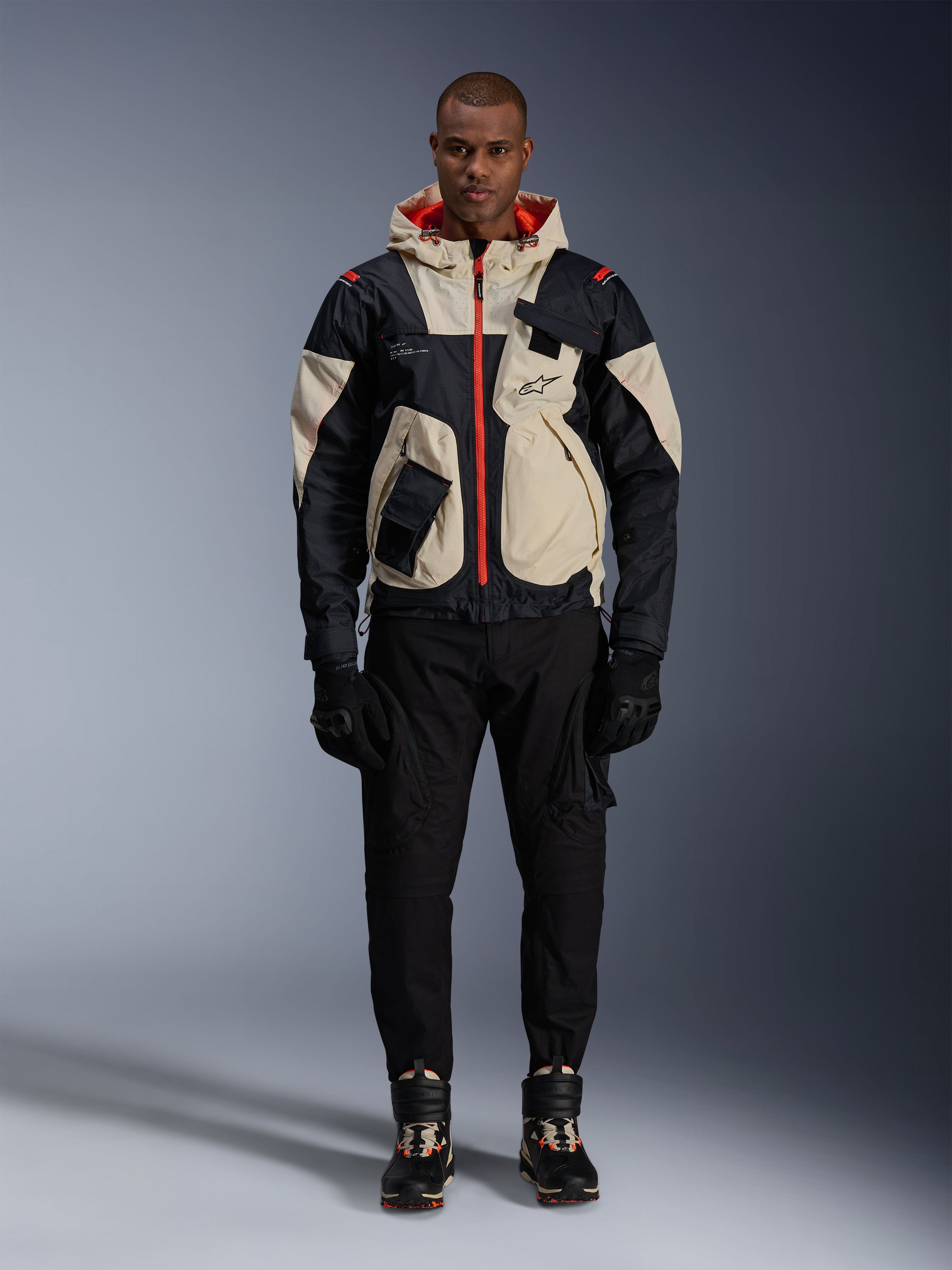 Alpinestars Mogress Air-jakke, urban motorcykeljakke, sort, Almond og Hot Orange, båret af en mandlig model med sorte bukser og handsker, utility-inspireret design med cremefarvede paneler og klare orange lynlåse, moderne motorsport-inspireret streetwear.