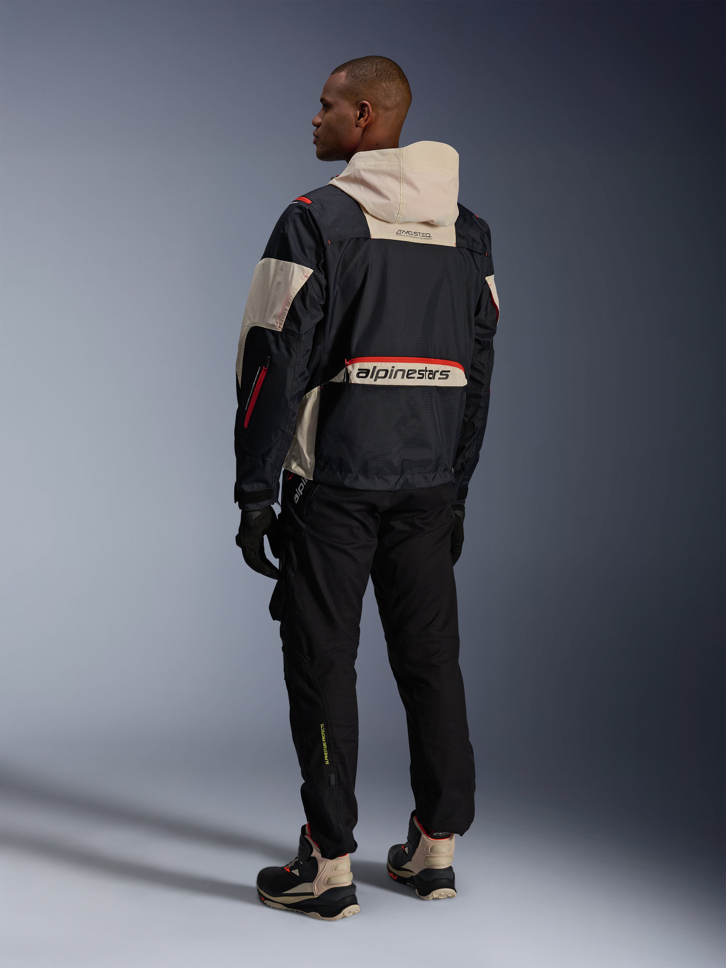 Set bagfra af en mand iført Alpinestars Acteon-bukser i sort, kombineret med en Mogress Air-jakke i sort og creme, sorte handsker og kørestøvler, moderne urbant motorcykeloutfit