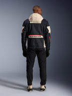 Set bagfra af en kører iført Alpinestars Mogress Air-jakke i Black, Almond og Hot Orange, med en cremefarvet hætte, stort Alpinestars-branding over baglommen og orange lynlåsdetaljer, kombineret med sorte bukser og cremefarvede støvler for et urbant motorcykel-look.