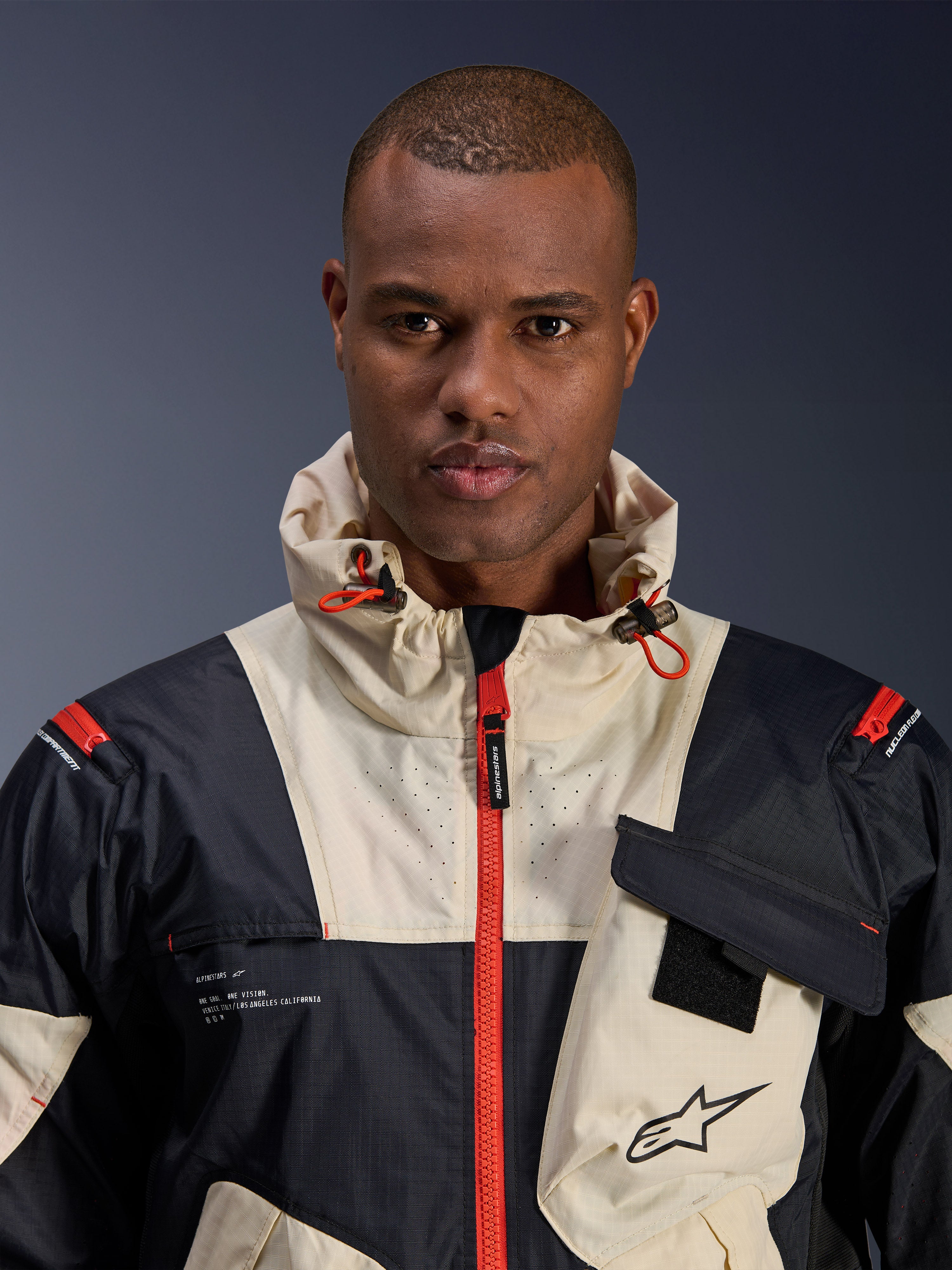Mandlig model iført Alpinestars Mogress Air-jakke, urban motorcykeljakke, Black Almond og Hot Orange, nærbillede der viser det farveblokerede design, ripstop-tekstur, hættekrave med orange stoppere og detaljer på frontlynlåsen.