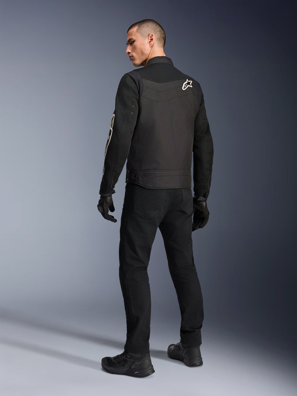 Alpinestars T-Dyno Air-jakke, sort, set bagfra af model iført en åndbar tekstil-motorcykeljakke, med hvidt Alpinestars-logo på skulderen, kombineret med sorte bukser og handsker, urban kørestil