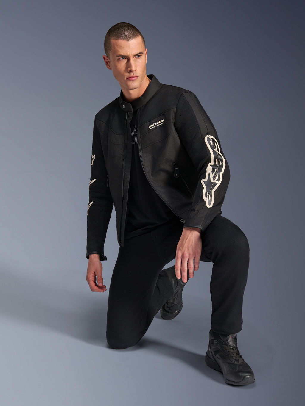 Mandlig model iført Alpinestars T-Dyno Air jakke, sort tekstil-motorcykeljakke med stor hvid branding på ærmerne, kombineret med sorte bukser og kørestøvler, moderne vintage-inspireret design med 3D-mesh-paneler.