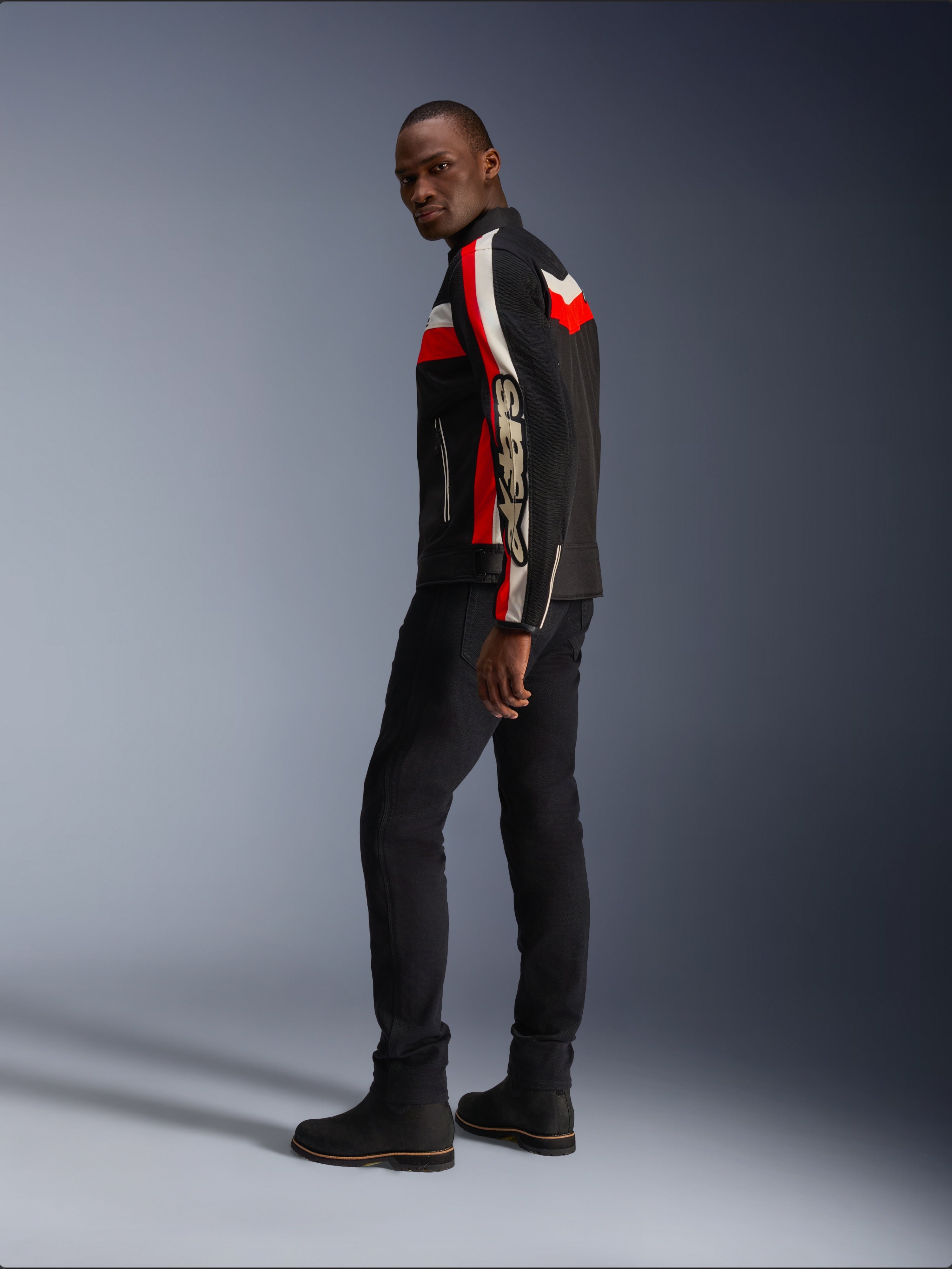 A man wearing the Alpinestars T-Dyno Air Jacket in Black, Bright Red, and Ecru, vintage-inspired textile motorcycle jacket with mesh panels, bold branding on the sleeve, paired with black pants and boots for a modern biker look