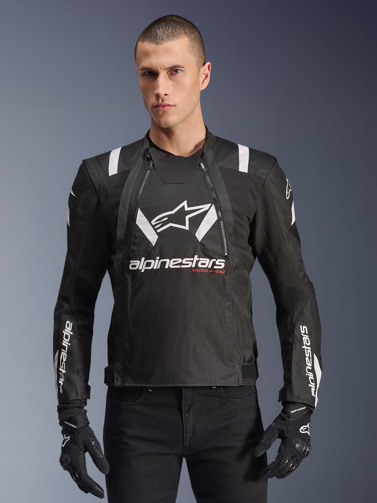 Alpinestars T-Stunt Air-jakke, sort og hvid motorcykeljakke, bæres af en mand med sorte handsker og bukser, med markant hvid branding på brystet og ærmerne og røde 'Racing Ahead'-detaljer, strømlinet sporty design med hvide skulderstriber og åndbare mesh-paneler.
