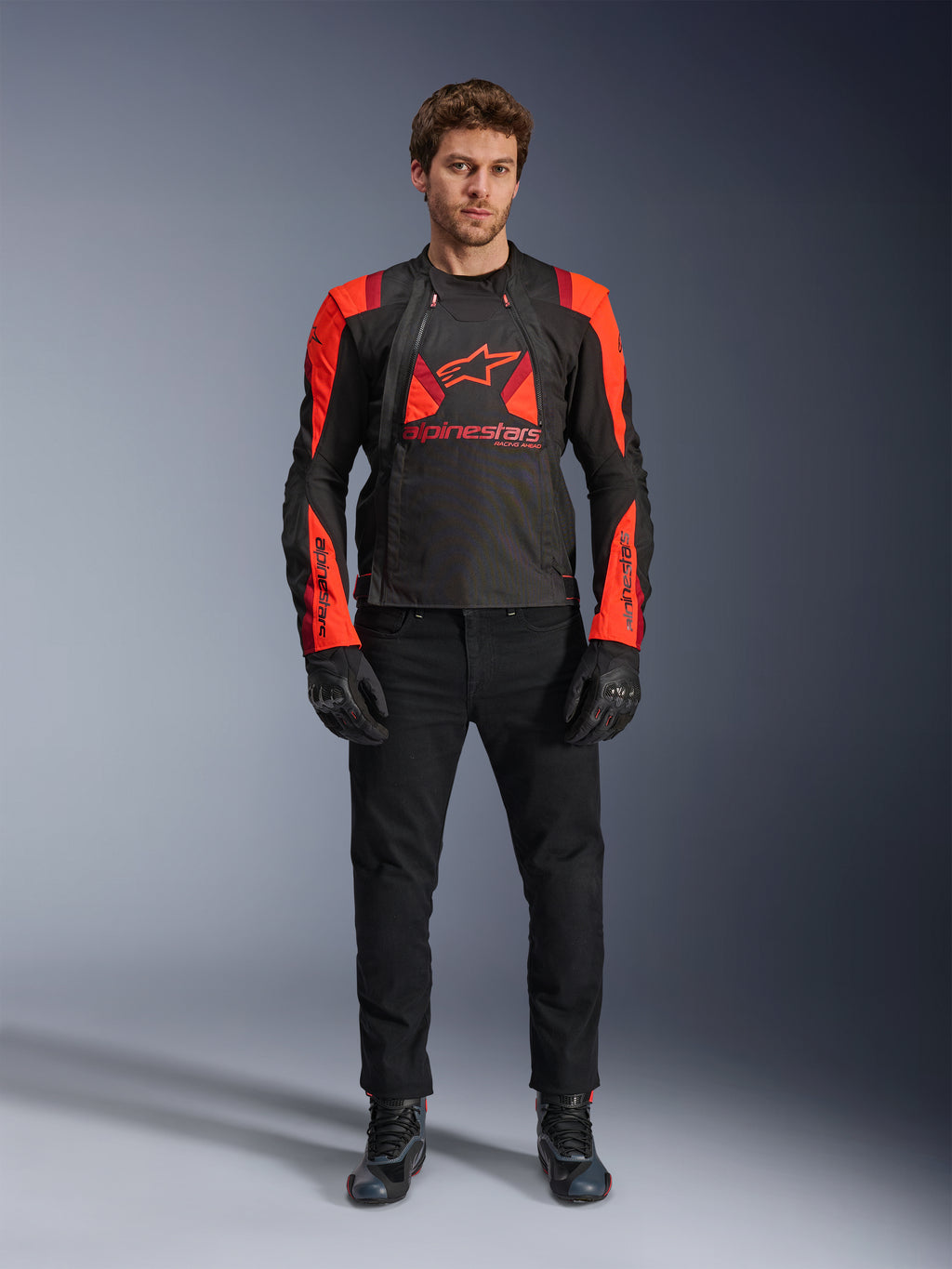 Alpinestars T-Stunt Air jakke, sort, fluo-rød og mørkerød, båret af en model med sorte kørejeans og handsker, med et modulært 2-i-1-design og store mesh-paneler for optimal luftgennemstrømning, motorcykeludstyr i sportssnit til landevejskørsel.