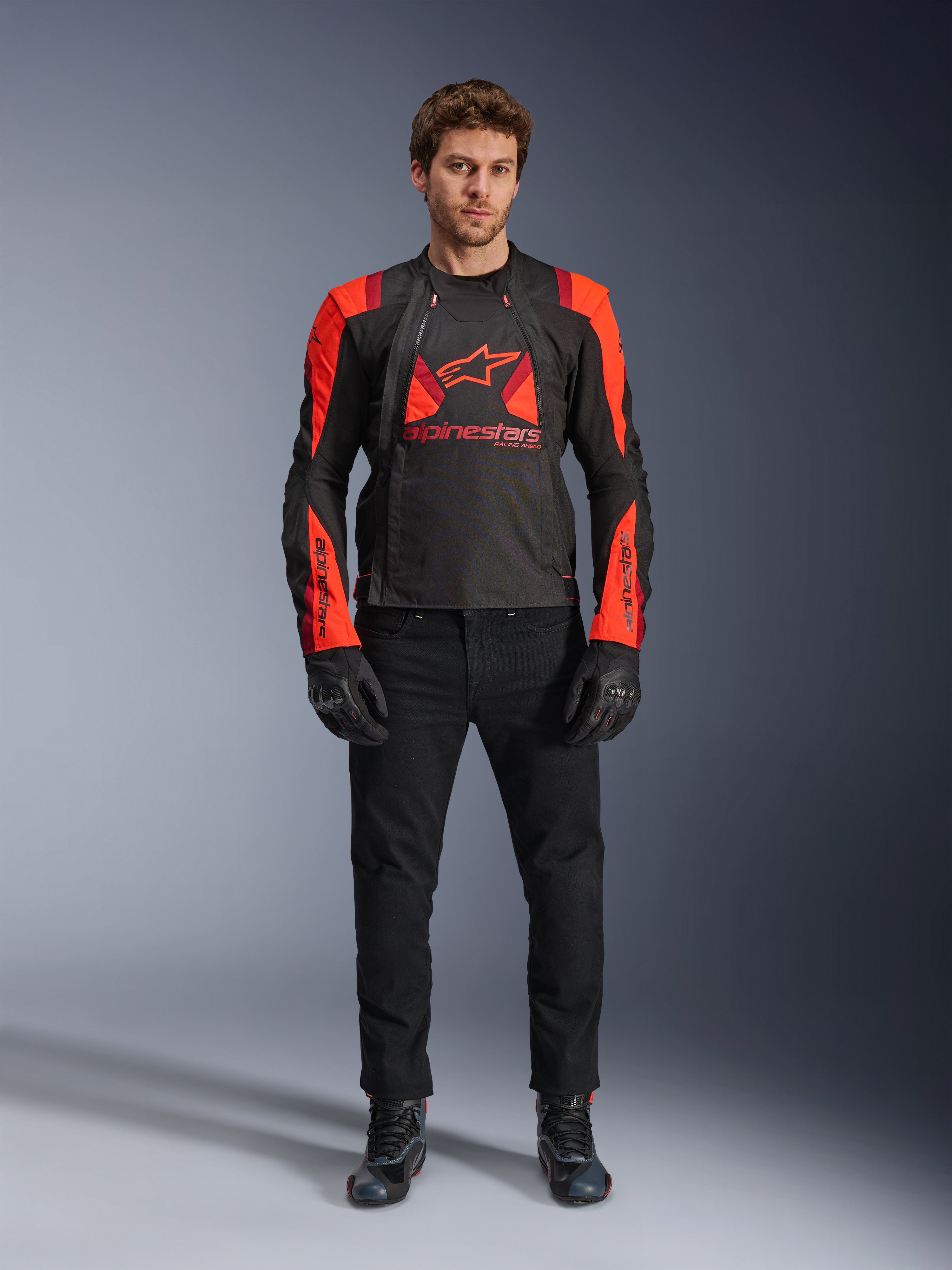 Alpinestars T-Stunt Air jakke, sort, fluo-rød og mørkerød, båret af en model med sorte kørejeans og handsker, med et modulært 2-i-1-design og store mesh-paneler for optimal luftgennemstrømning, motorcykeludstyr i sportssnit til landevejskørsel.