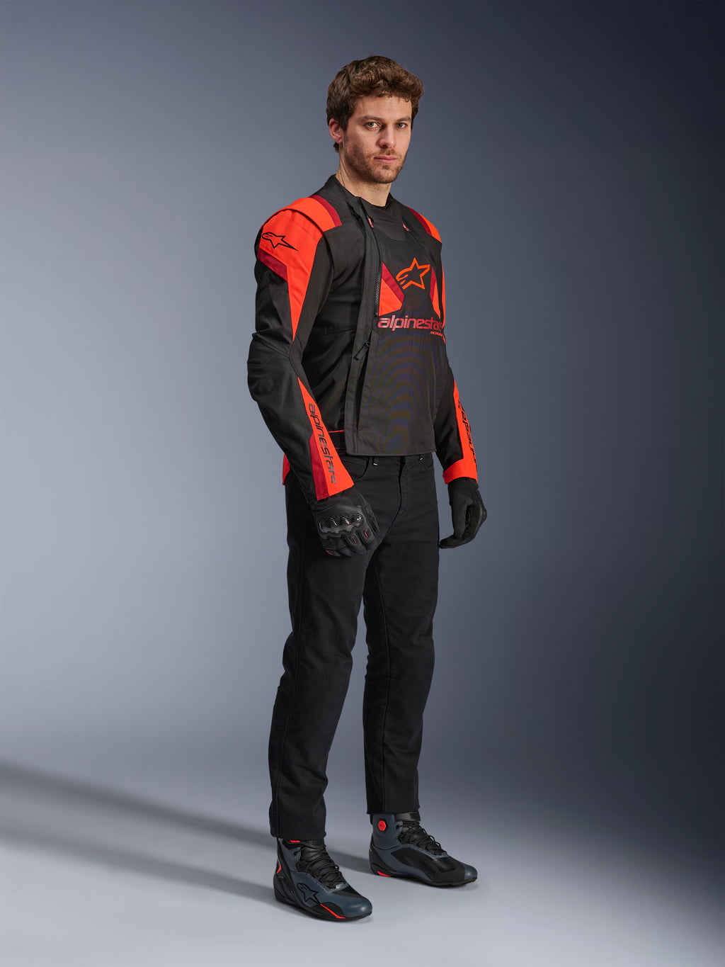 Alpinestars T-Stunt Air jakke, sort, rød fluo og mørkerød, modulær motorcykeljakke båret af en model med frontpanelerne åbne for at afsløre mesh-interiøret, kombineret med sorte motorcykelbukser, handsker og sko.