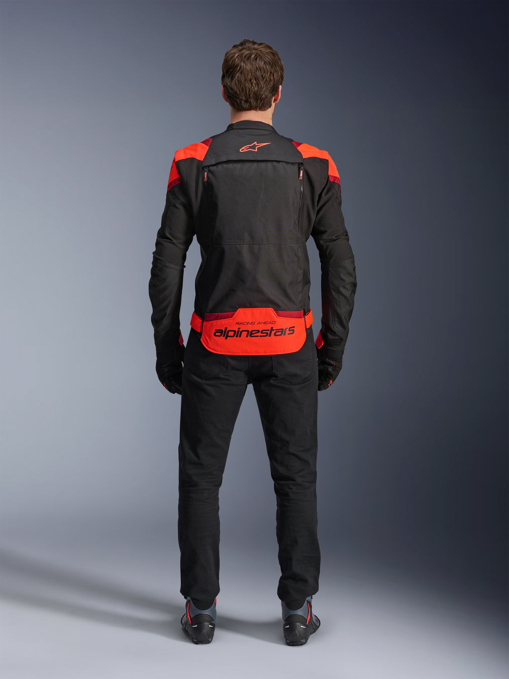 Alpinestars T-Stunt Air Jacket, sort med Red Fluo og mørkerøde detaljer, set bagfra på model, kombineret med sorte kørebukser og støvler, markant Alpinestars-branding på det nederste rygpanel, modulært design med mesh-konstruktion for ventilation.