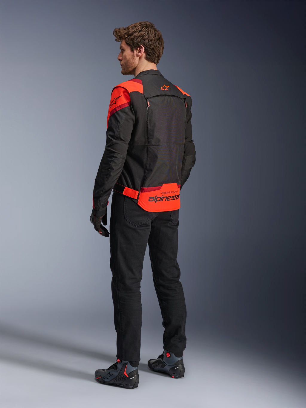 Mand iført Alpinestars T-Stunt Air-jakke set bagfra, sort, rød fluo og mørkerød, tekstil motorcykeljakke med mesh-paneler og sportsdesign, med tydeligt Alpinestars-branding på lænden, stylet med sorte bukser og kørestøvler