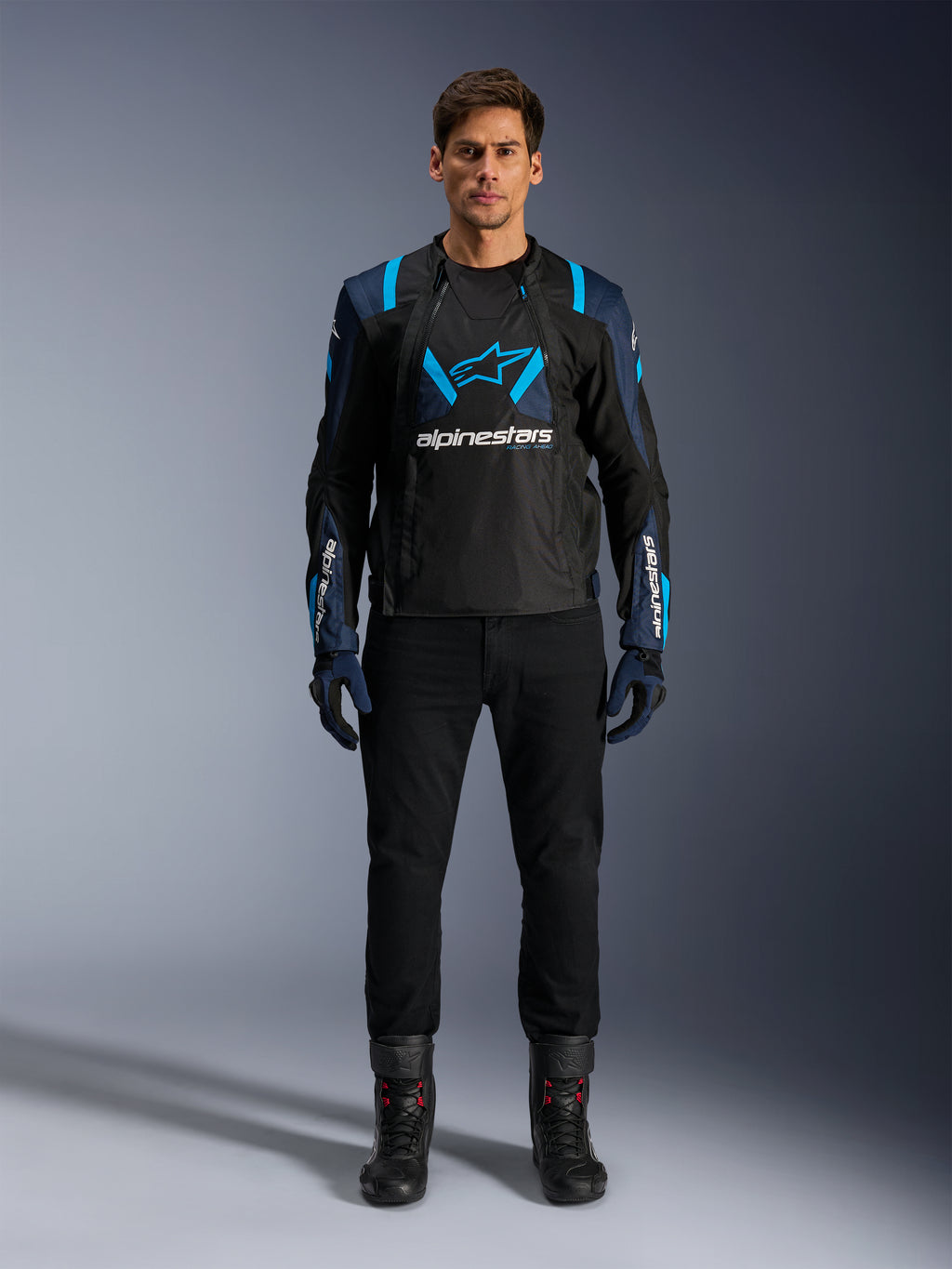 Alpinestars T-Stunt Air-jakke, motorcykeljakke, sort med mørkeblå og lyseblå detaljer, med et stort hvidt logo på brystet, mesh-konstruktion i sportssnit for luftgennemstrømning, båret med sorte kørebukser og støvler for en stilfuld æstetik til landevejskørsel.