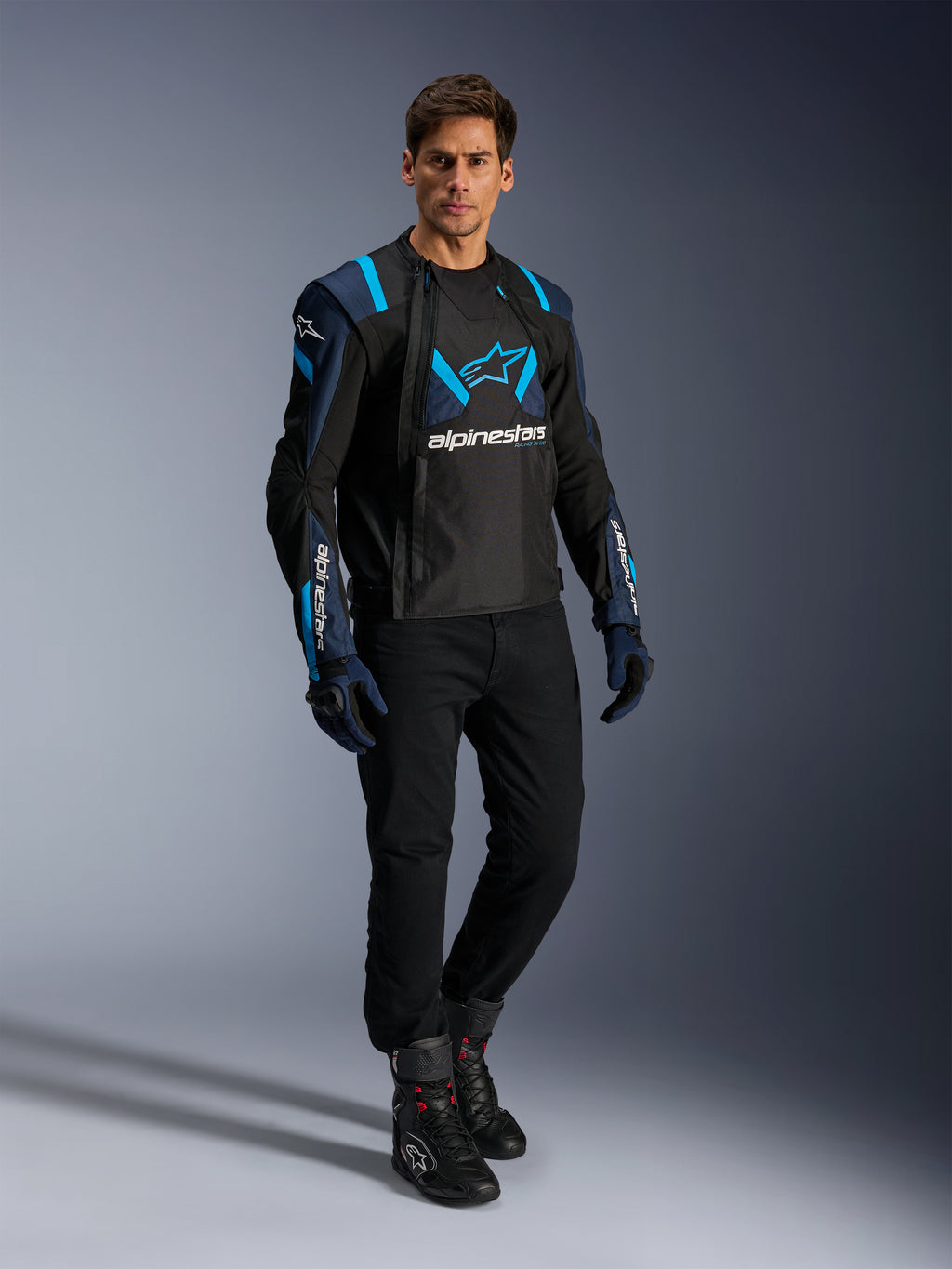 Mandlig model iført Alpinestars T-Stunt Air jakke, sort, mørkeblå og lyseblå, med fremtrædende mesh-paneler for luftgennemstrømning, kombineret med sorte bukser og kørehandsker, sportslig motorcykeljakke til sommerens landevejskørsel.