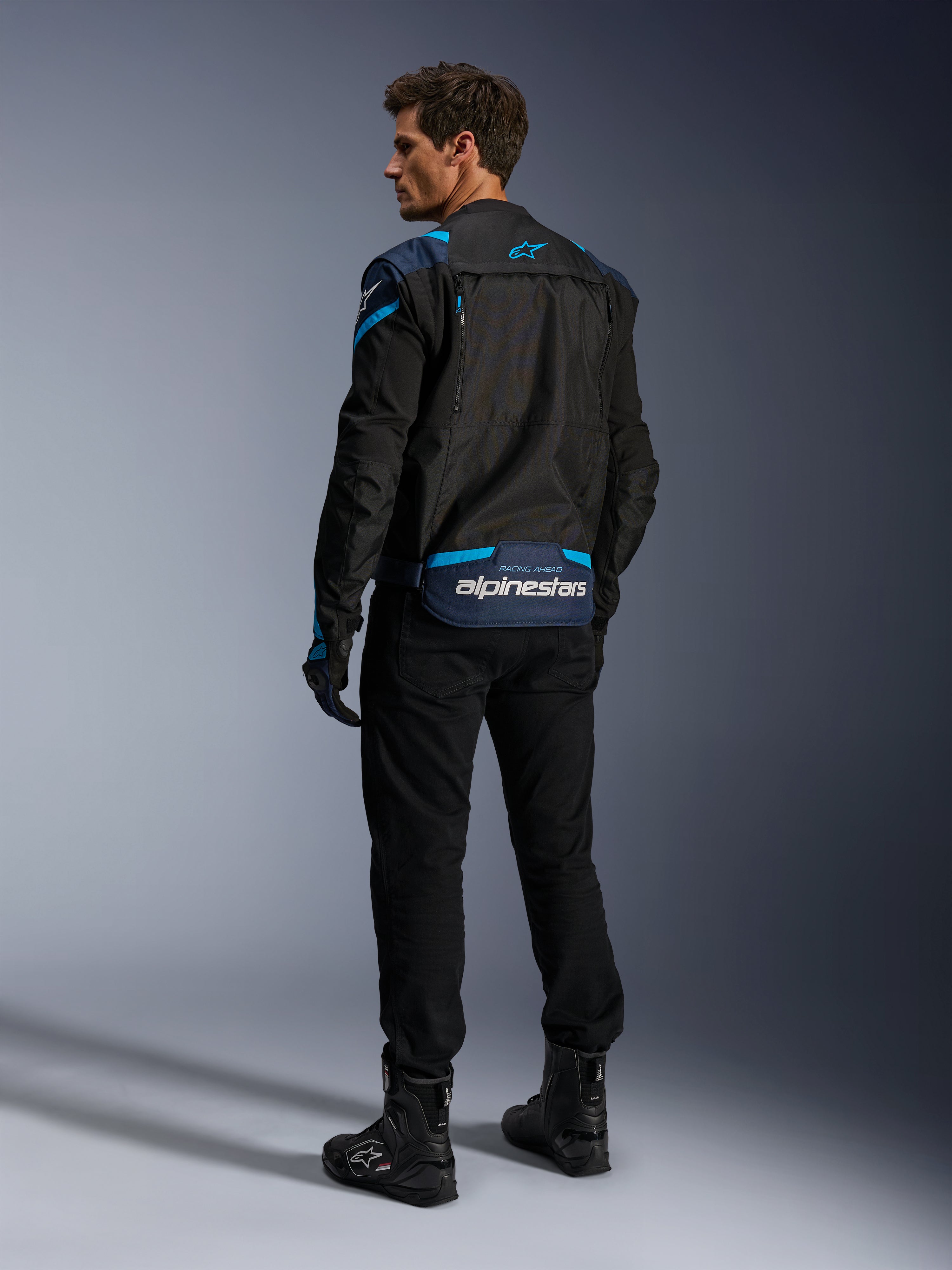 Alpinestars T-Stunt Air jakke, motorcykeljakke, sort med mørkeblå og lyseblå detaljer, set bagfra med modulære mesh-paneler og branding på lænden, båret af en model med sorte kørebukser og støvler