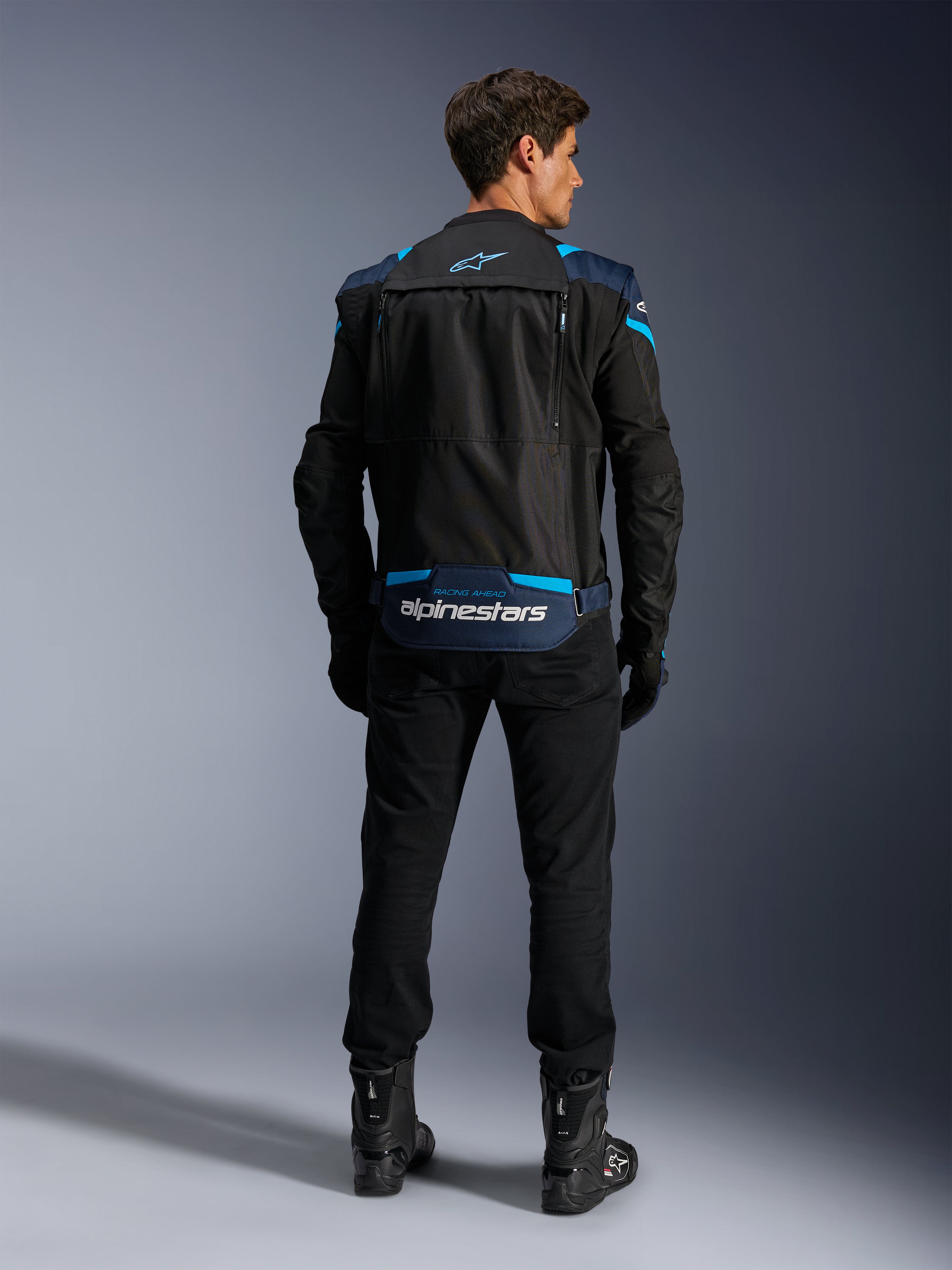 Alpinestars T-Stunt Air-jakke, modulær tekstil-motorcykeljakke, sort, mørkeblå og lyseblå, set bagfra, der viser det nederste opbevaringsrum til ærmer med hvidt Alpinestars-branding, båret med sorte bukser og støvler.