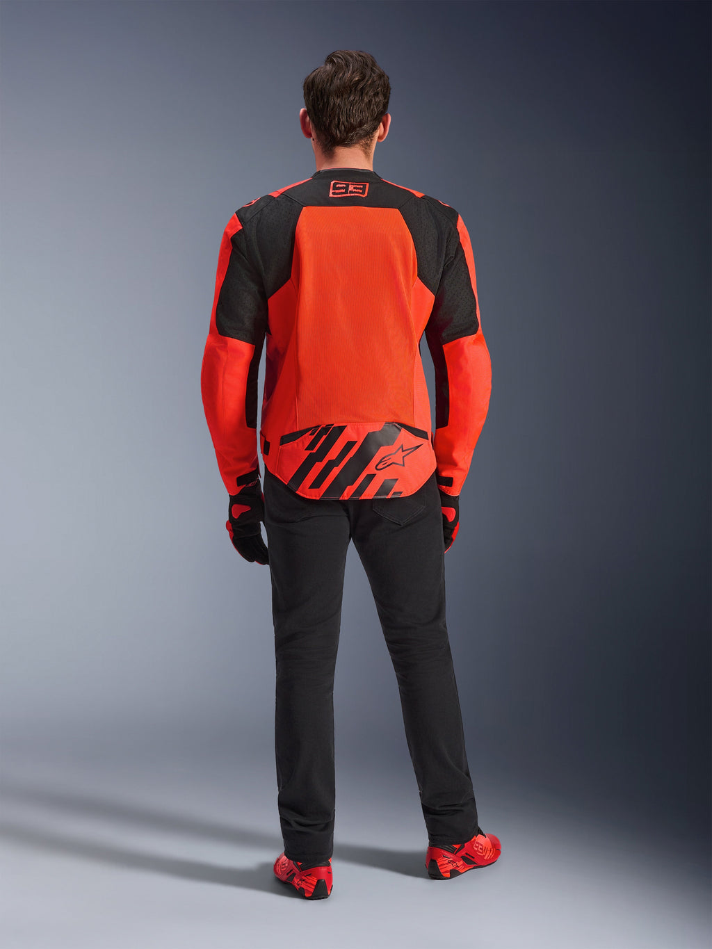 Set bagfra af MM93 T-SPS Air V2-jakken, neonrød og sort, tekstil motorcykeljakke med store mesh-paneler på ryggen til ventilation, båret med sorte bukser og rødt fodtøj, med Marc Marquez 93-branding