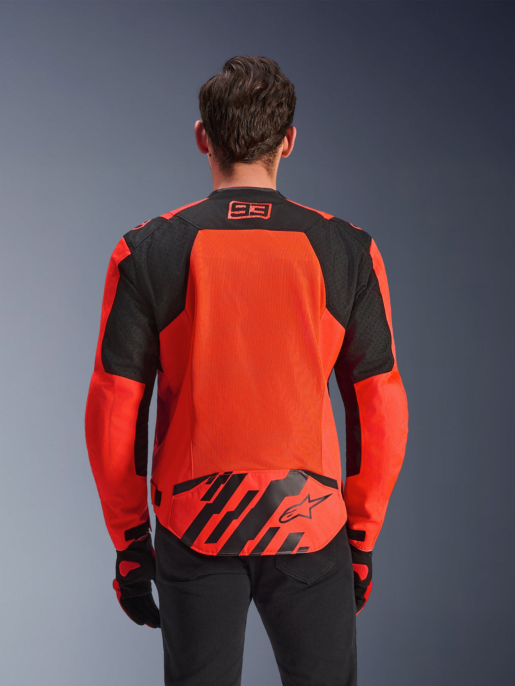 Alpinestars MM93 T-SPS Air V2 jakke, set bagfra, rød fluo og sort, med 93-racinglogo i nakken, stort mesh-panel på ryggen for luftgennemstrømning, flap nederst på ryggen med Alpinestars-stjernelogo og geometriske striber, vist med sorte bukser og kørehandsker.