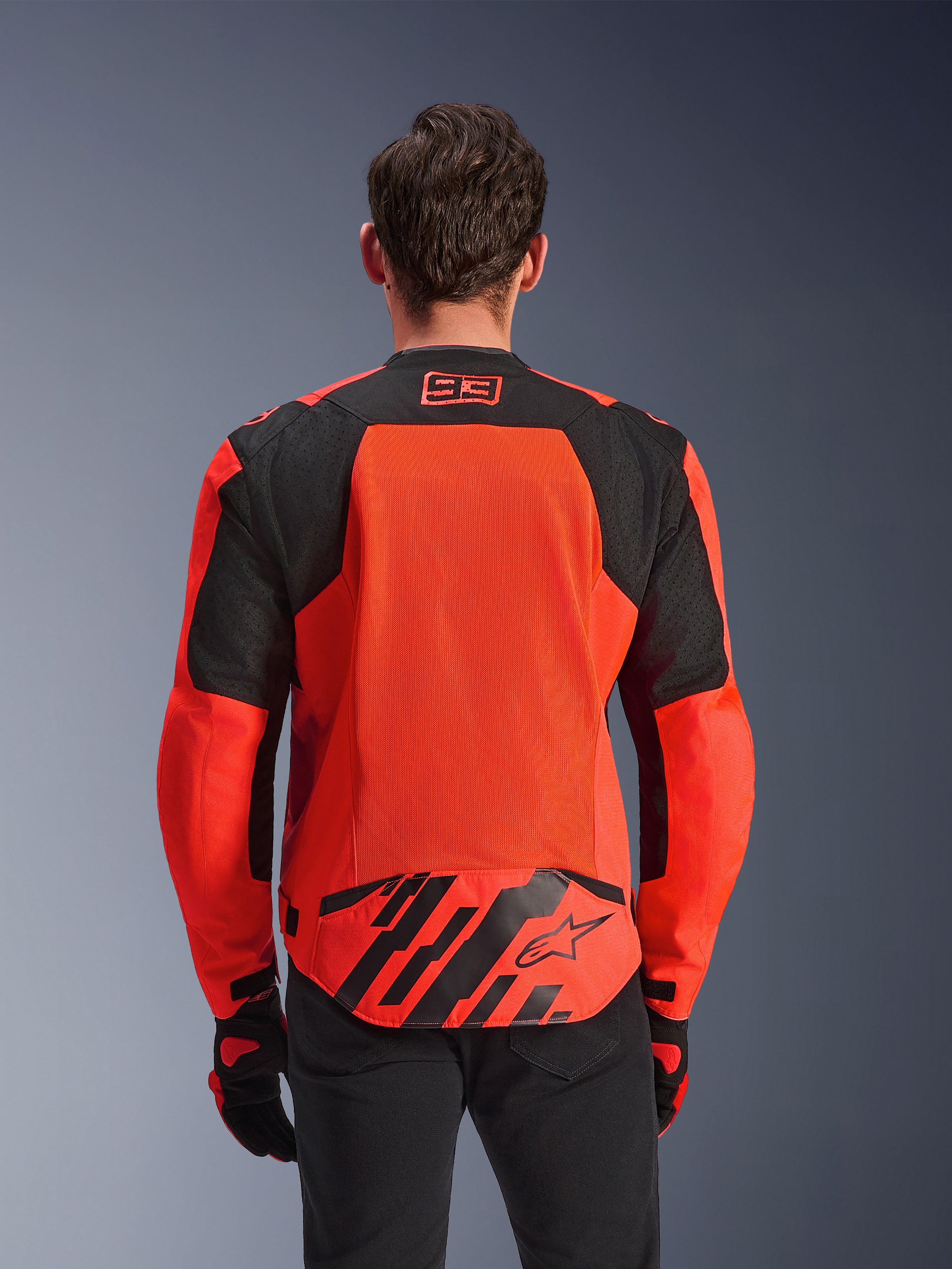 Alpinestars MM93 T-SPS Air V2 jakke, set bagfra, rød fluo og sort, med 93-racinglogo i nakken, stort mesh-panel på ryggen for luftgennemstrømning, flap nederst på ryggen med Alpinestars-stjernelogo og geometriske striber, vist med sorte bukser og kørehandsker.