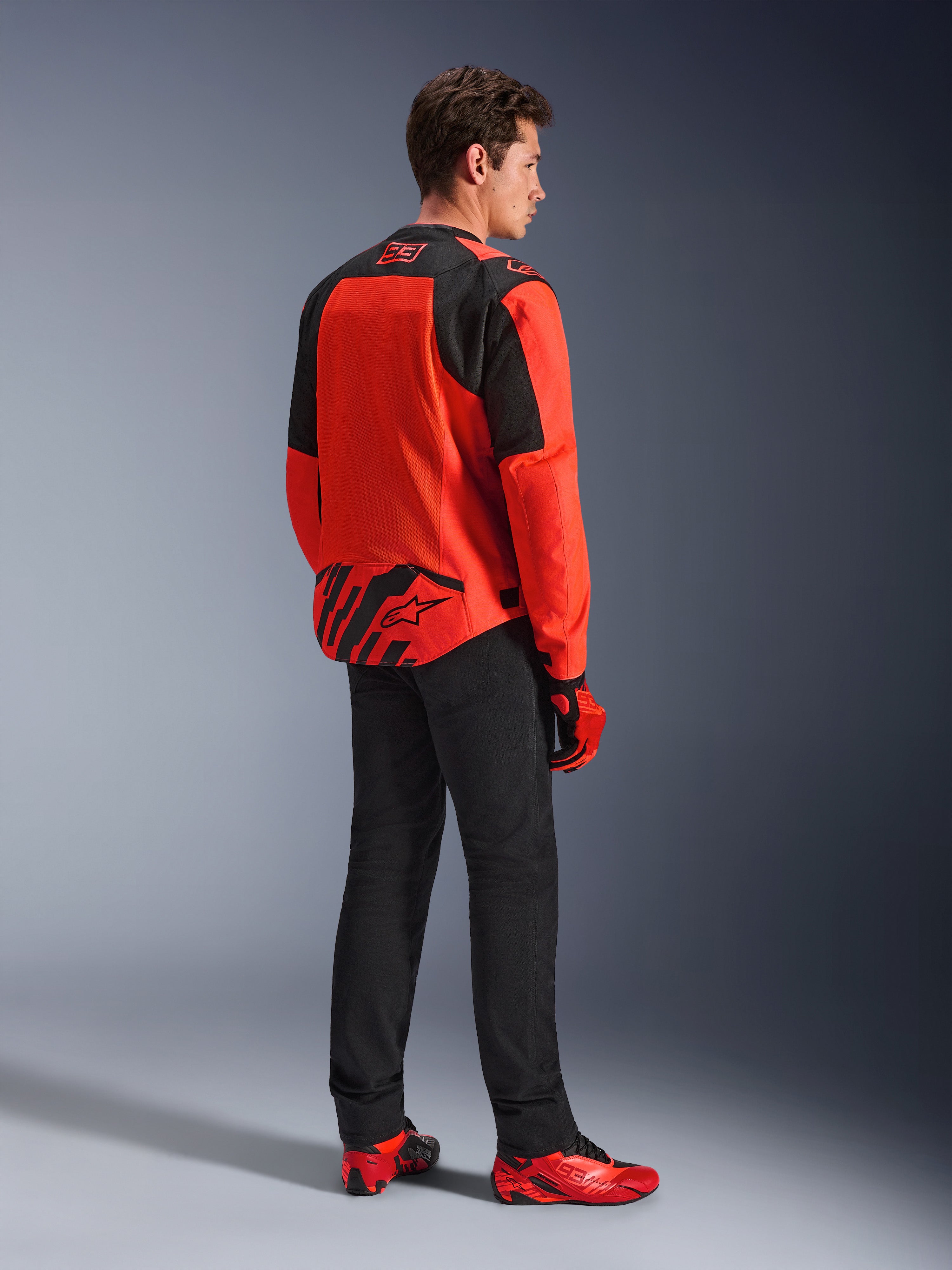 Alpinestars MM93 T-SPS Air V2 jakke i rød fluo og sort, trekvartvisning bagfra på en model, med Marc Marquez 93-branding på kraven og den nederste del af ryggen, stylet med sorte bukser og røde motorcykelsko.