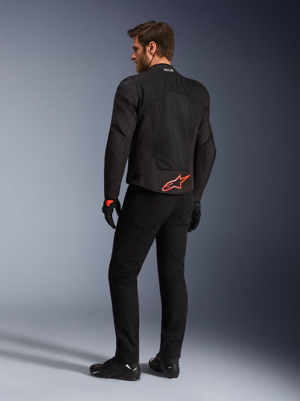 Alpinestars SMX Air-jakke i sort, set bagfra på en model, med et stærkt rødt Alpinestars-logo på lænden, 'Tech-Air Ready'-branding på kraven, tekniske mesh-paneler til ventilation, kombineret med sorte kørebukser og handsker.