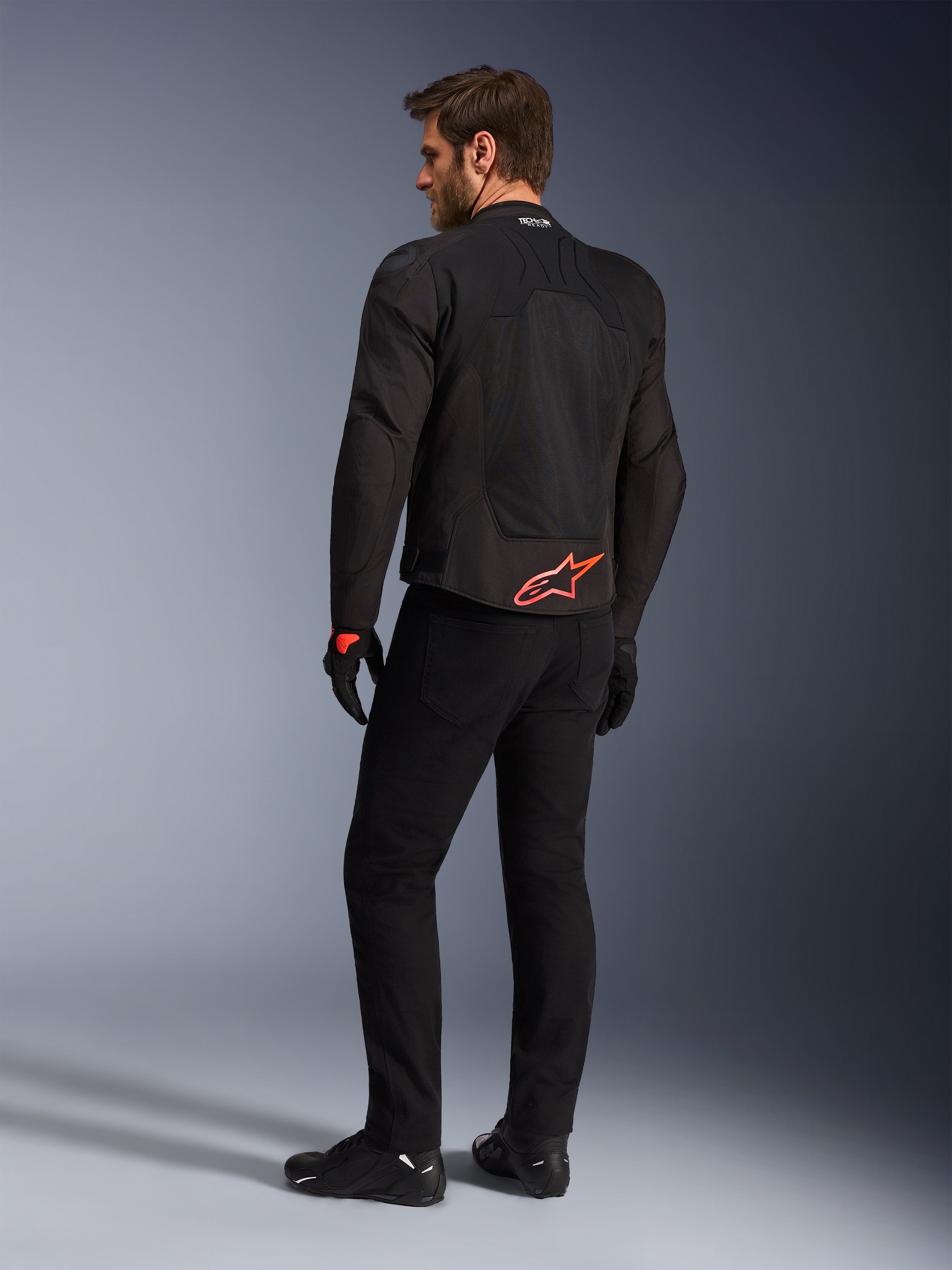 Alpinestars SMX Air-jakke i sort, set bagfra på en model, med et stærkt rødt Alpinestars-logo på lænden, 'Tech-Air Ready'-branding på kraven, tekniske mesh-paneler til ventilation, kombineret med sorte kørebukser og handsker.