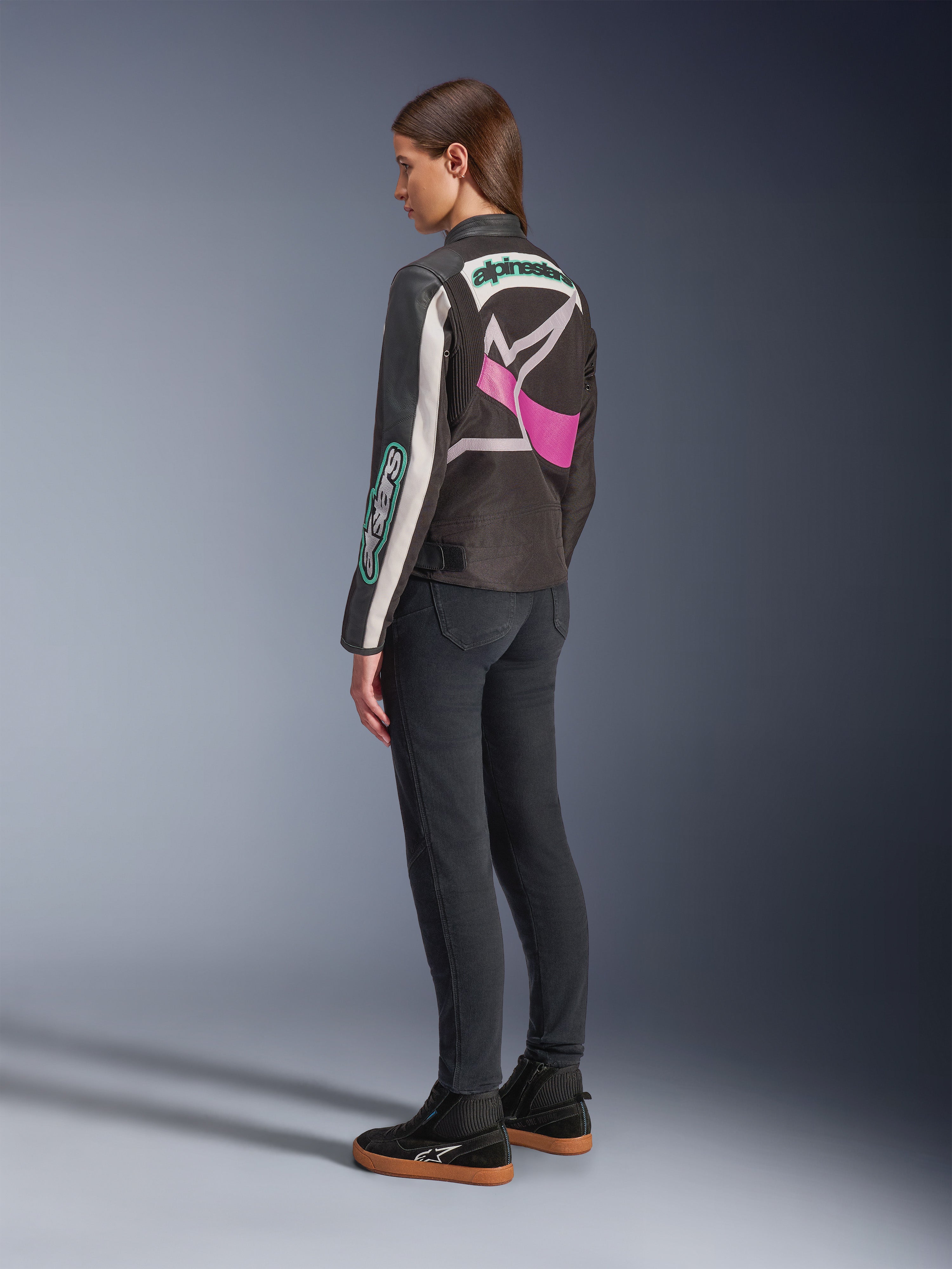 Alpinestars Idles Women’s Jacket, Motorcycle Jacket, Black Off White and Purple, viewed from the back on model, featuring a large wrap-around Alpinestars logo and vintage racing-inspired design, paired with dark jeans and black shoes.