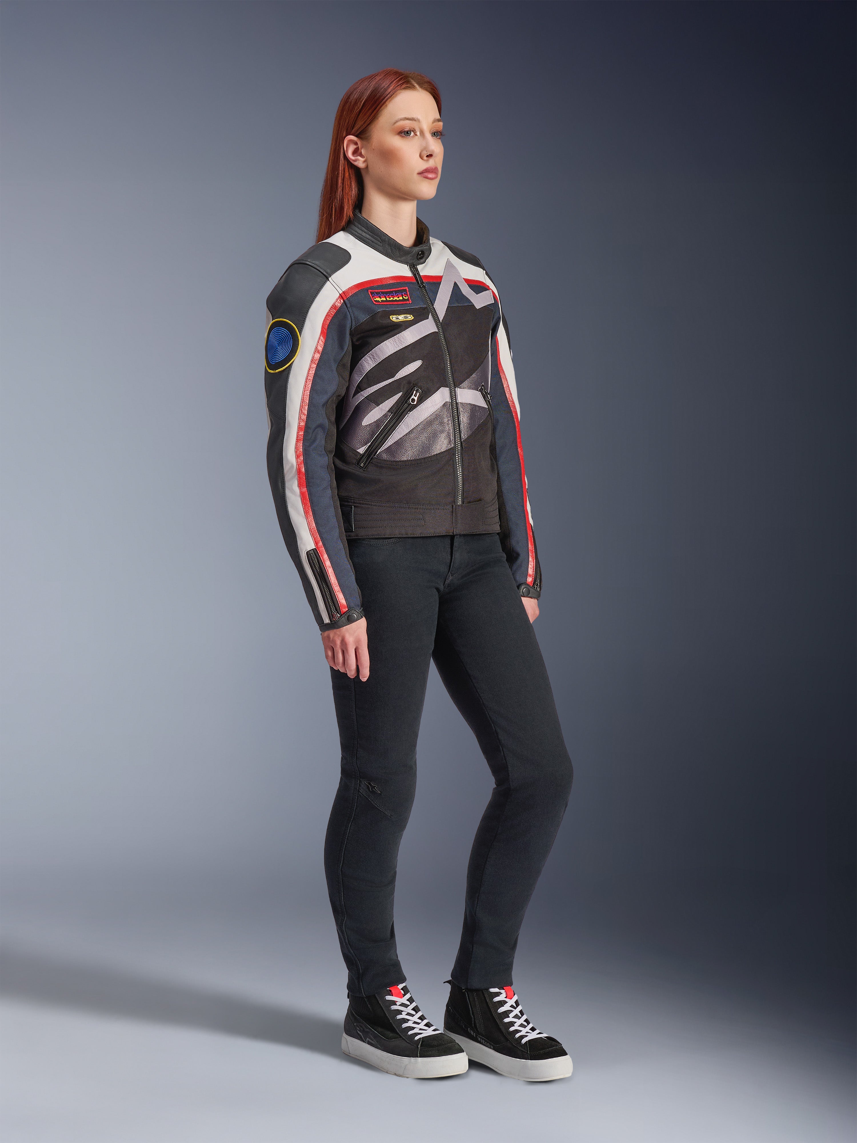 Alpinestars Idles damejakke, motorcykeljakke, off-white, flammerød og sort, med et stort kropsomsluttende Alpinestars-logo og vintage racing-inspirerede patches, båret med sorte bukser og high-top sneakers, moderne urbant køreudstyr