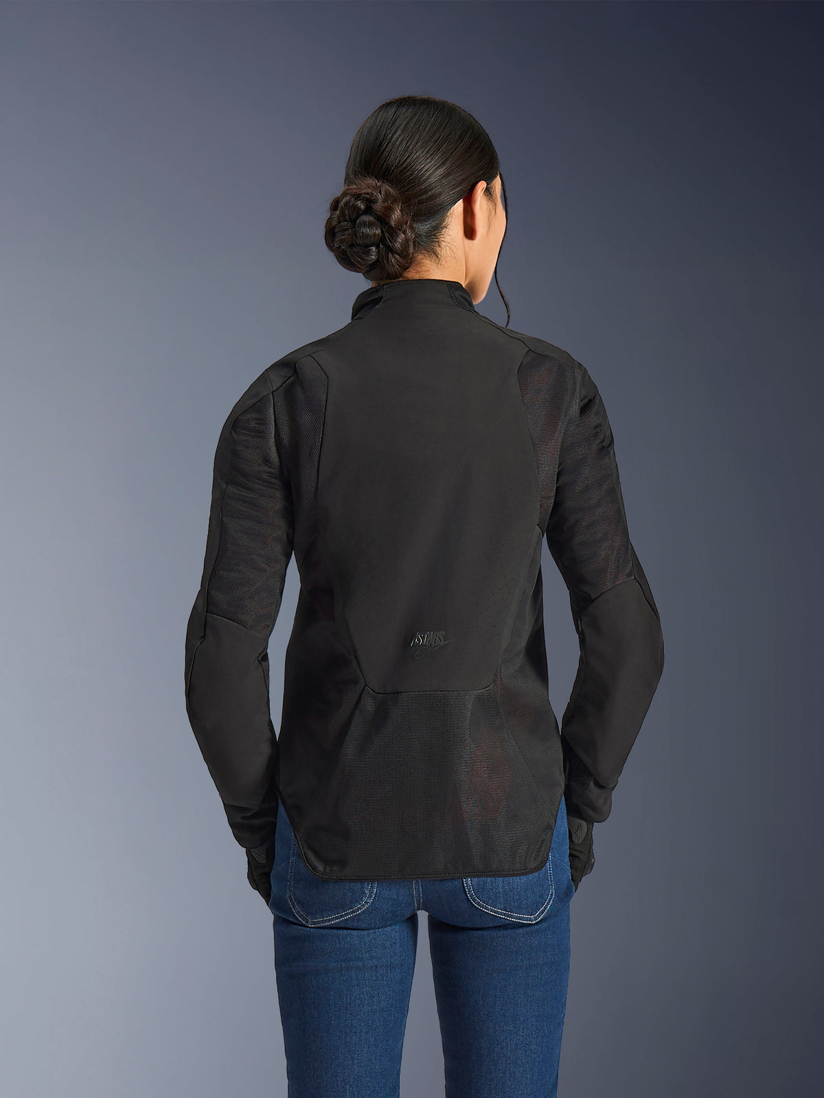 Alpinestars Woman Stella Aeroshell Airflow-jakke, Black Anthracite, set bagfra med integrerede mesh-ventilationspaneler på ryg og ærmer, laserperforeret softshell-chassis med forlænget rygprofil, båret med blå denimjeans.