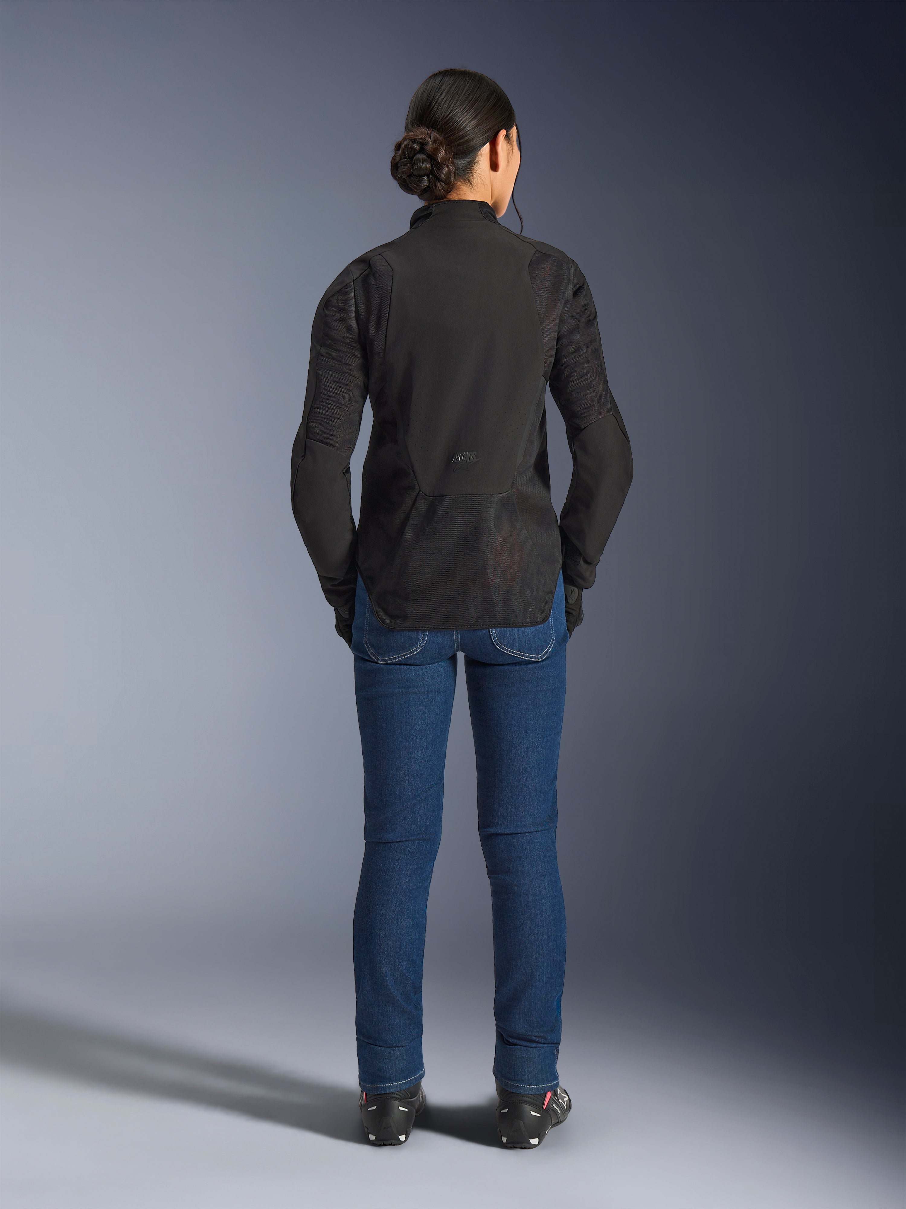 Alpinestars Stella Aeroshell Airflow jakke til kvinder, Black Anthracite, set bagfra på en model, letvægts softshell-chassis med laserperforerede mesh-paneler, forlænget bagprofil for dækning, sportslig pasform, kombineret med blå jeans