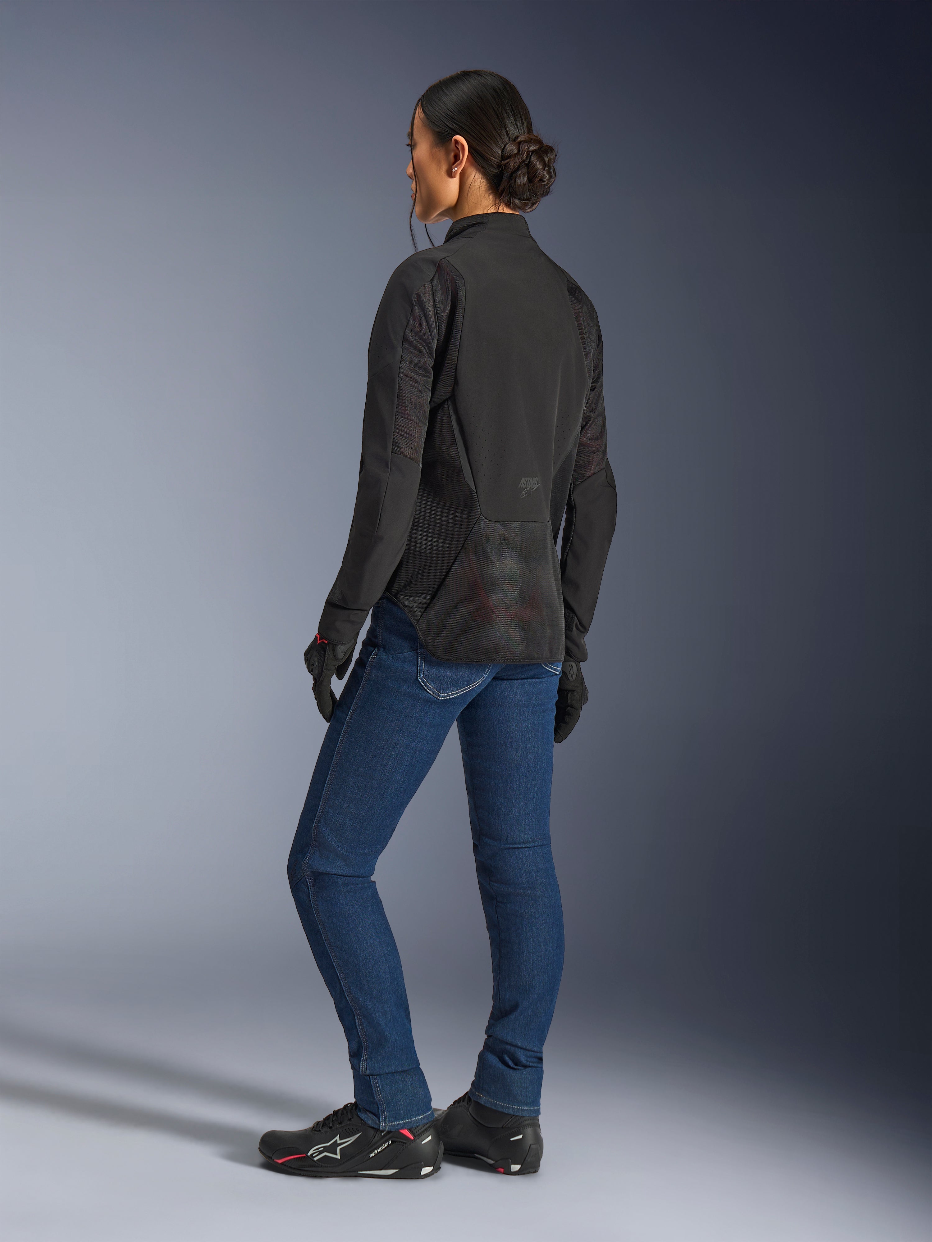 Kvinde iført Alpinestars Stella Aeroshell Airflow-jakke, Black Anthracite, trekvartvisning bagfra, der viser åndbare mesh-paneler og en forlænget bagprofil, kombineret med blå denim motorcykeljeans og sorte Alpinestars-sko