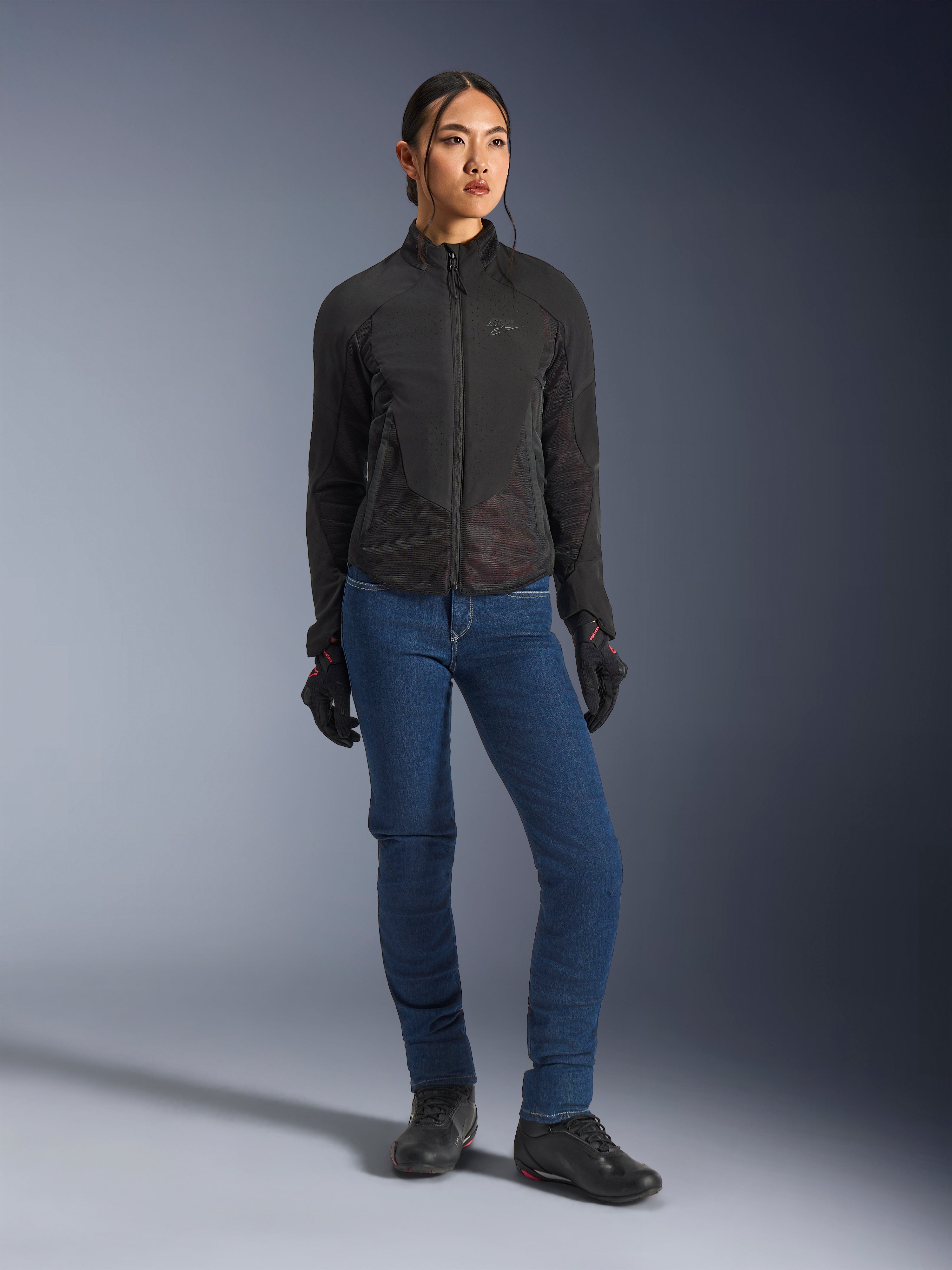 Kvindelig rytter iført Alpinestars Stella Aeroshell Airflow-jakken i sort, med laserperforeret ventilation og mesh-paneler, stylet med blå motorcykeljeans og sort motorcykeludstyr, moderne sportslig og urban køreæstetik