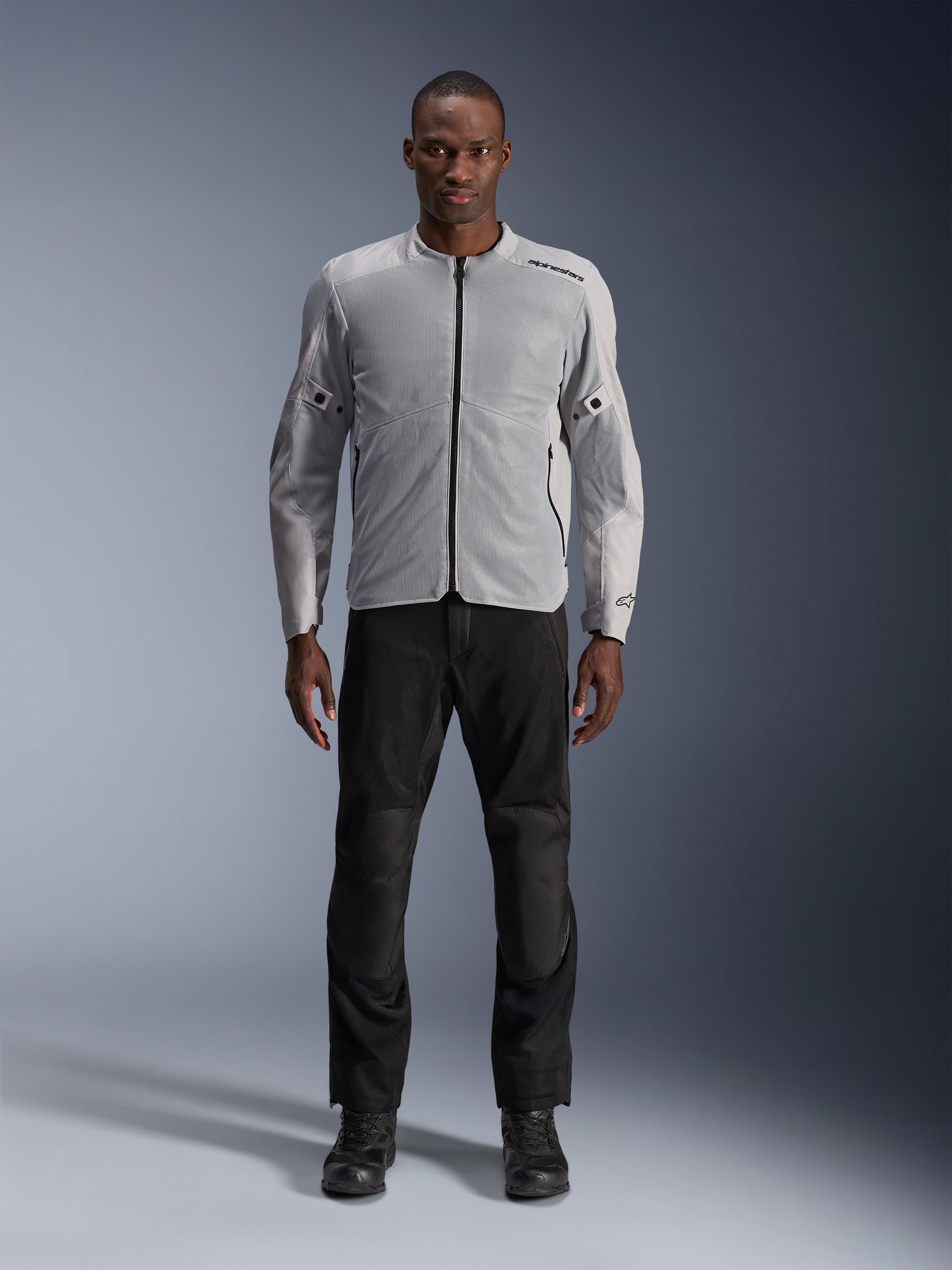 Alpinestars C-1 Air-jakke i sølv, urban tekstil-motorcykeljakke til mænd med ventilerede mesh-paneler, båret af en model med sorte kørebukser og sorte støvler, moderne pendlingsstil