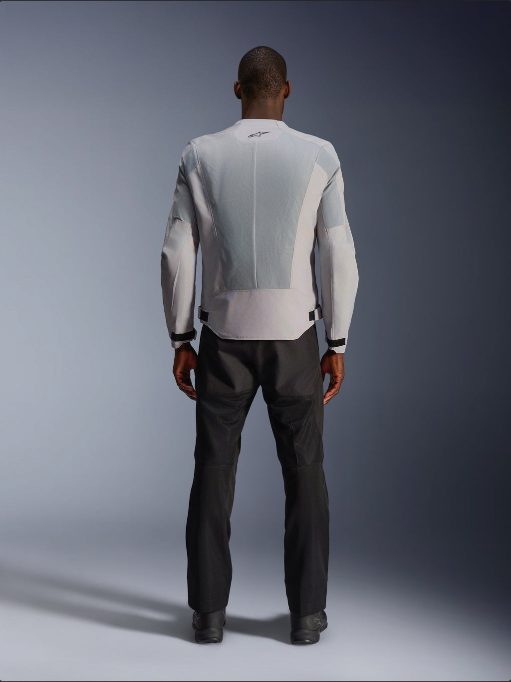 Alpinestars C-1 Air-jakke i sølv, set bagfra på en mandlig model, tekstil motorcykeljakke med store tech mesh-ventilationspaneler, lille sort logo i nakken og taljejusteringer, båret med sorte kørebukser for et moderne urbant look.