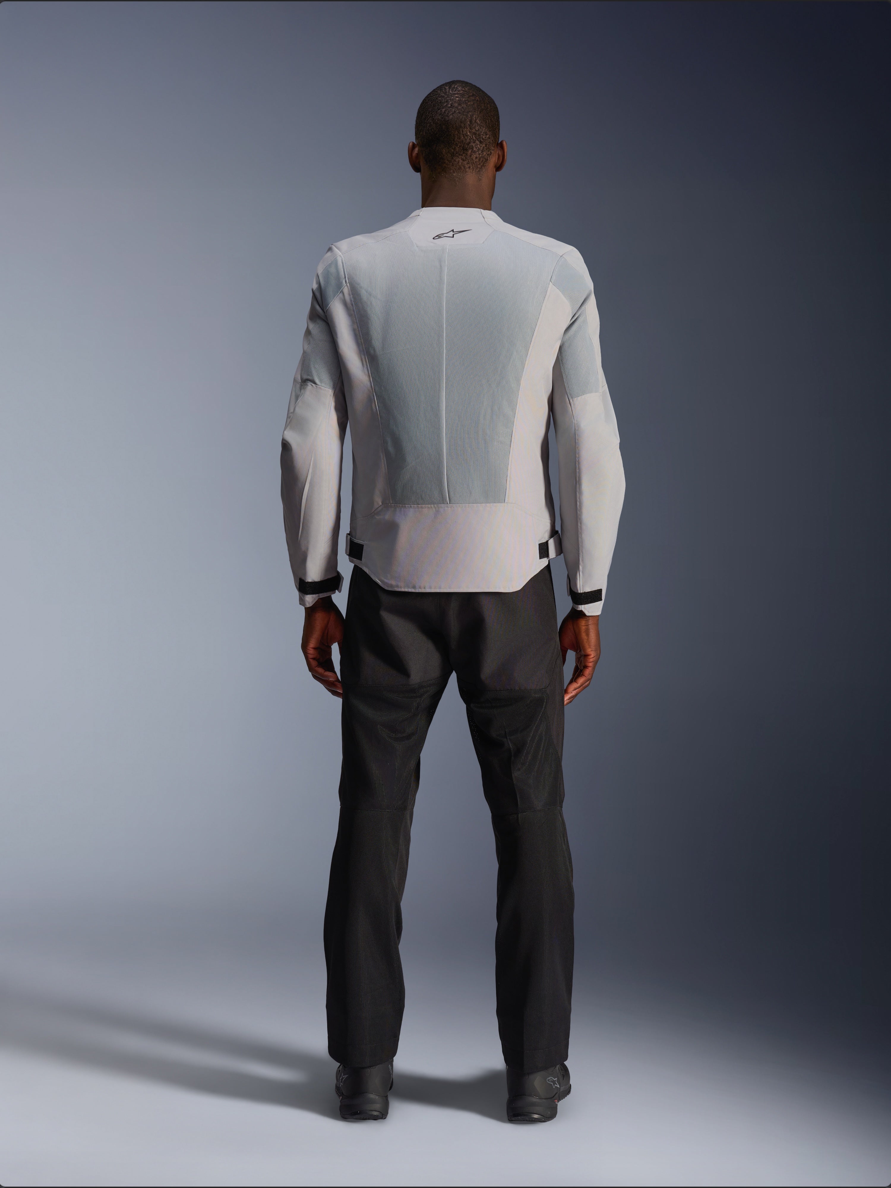 Alpinestars C-1 Air-jakke i sølv, set bagfra på en mandlig model, tekstil motorcykeljakke med store tech mesh-ventilationspaneler, lille sort logo i nakken og taljejusteringer, båret med sorte kørebukser for et moderne urbant look.