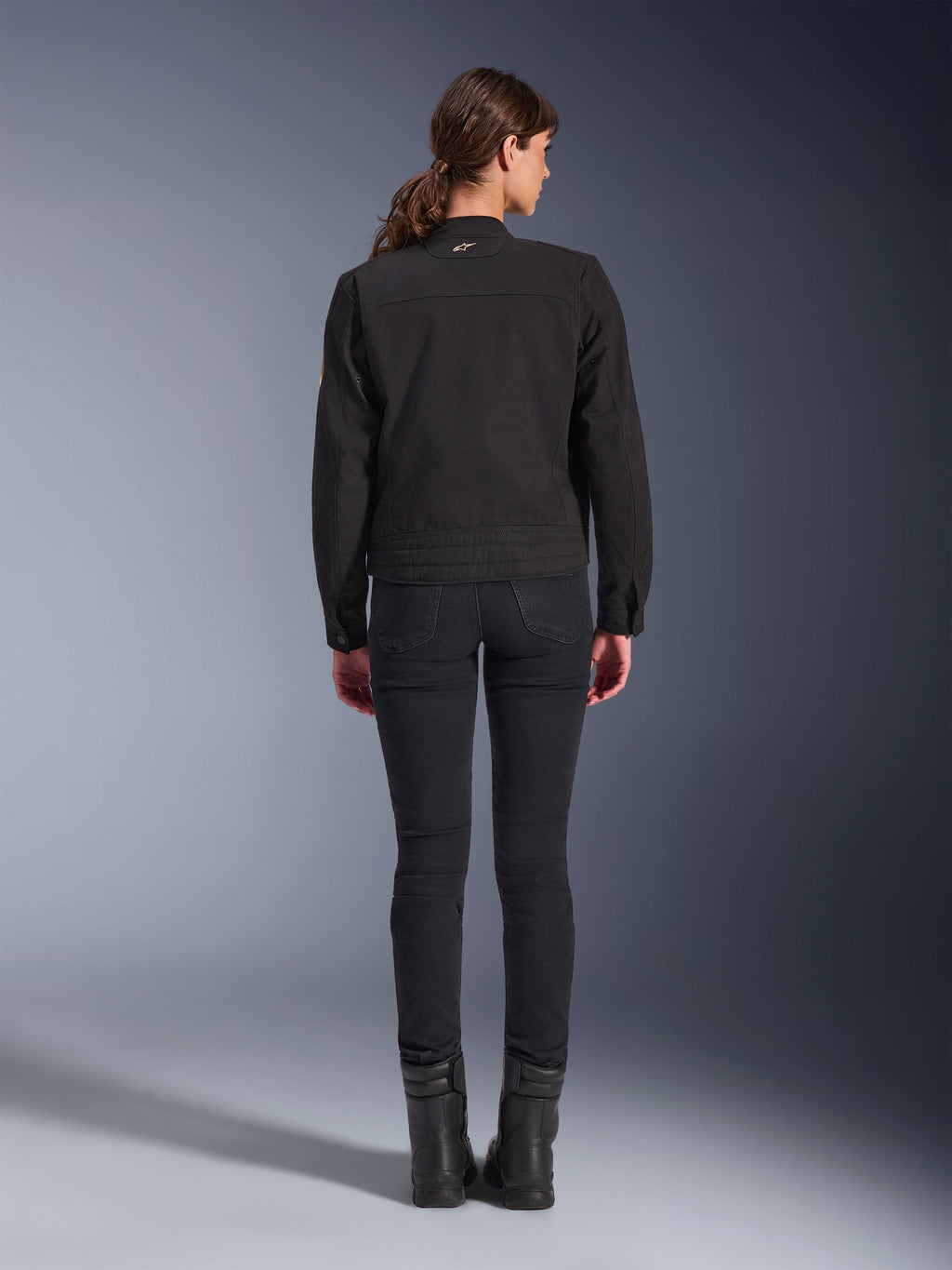 Alpinestars Stella Grace-jakke, urban motorcykeljakke til kvinder, sort, set bagfra der viser det arbejdstøjsinspirerede design, båret med sorte slim-fit bukser og sorte støvler, studiebillede