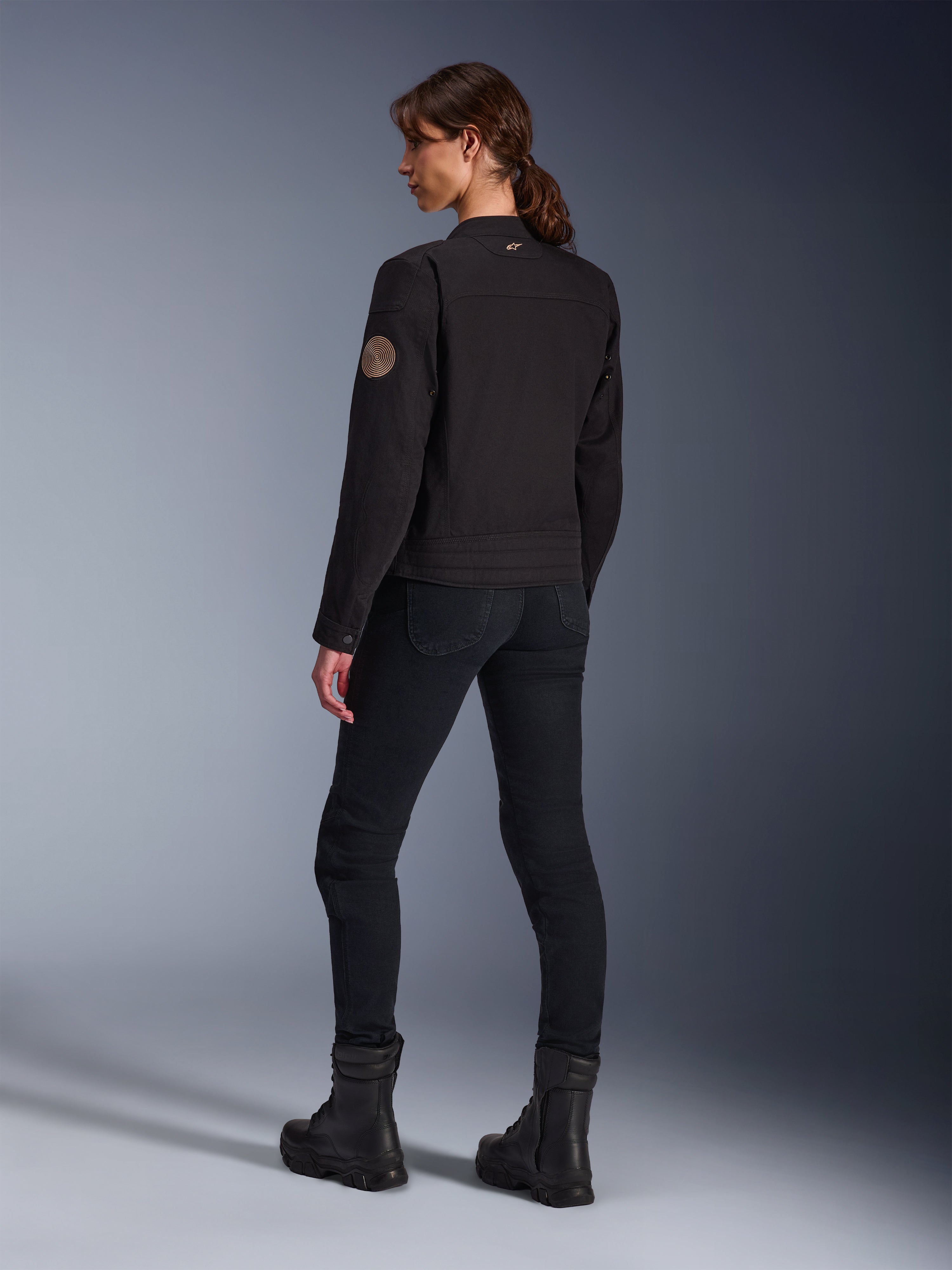 Alpinestars Stella Grace-jakke i sort, model set bagfra, der fremhæver vintage Oscar-spiralmærket på armen og det lille stjernelogo på kraven, kombineret med sorte motorcykelbukser og støvler til bykørsel