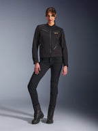 Kvinde iført Alpinestars Stella Grace-jakke i sort, urban motorcykeljakke med arbejdstøjs-æstetik, med lynlåslommer foran og brystlogo, kombineret med sorte slim-fit bukser og sorte støvler, moderne køreudstyr til kvinder.