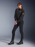 Kvindelig model iført Alpinestars Stella Grace jakke til kvinder, sort urban motorcykeljakke, stylet med sorte kørebukser og støvler, arbejdstøjsinspireret design med bronzefarvet broderet logo på brystet og et rundt spiral-patch på ærmet