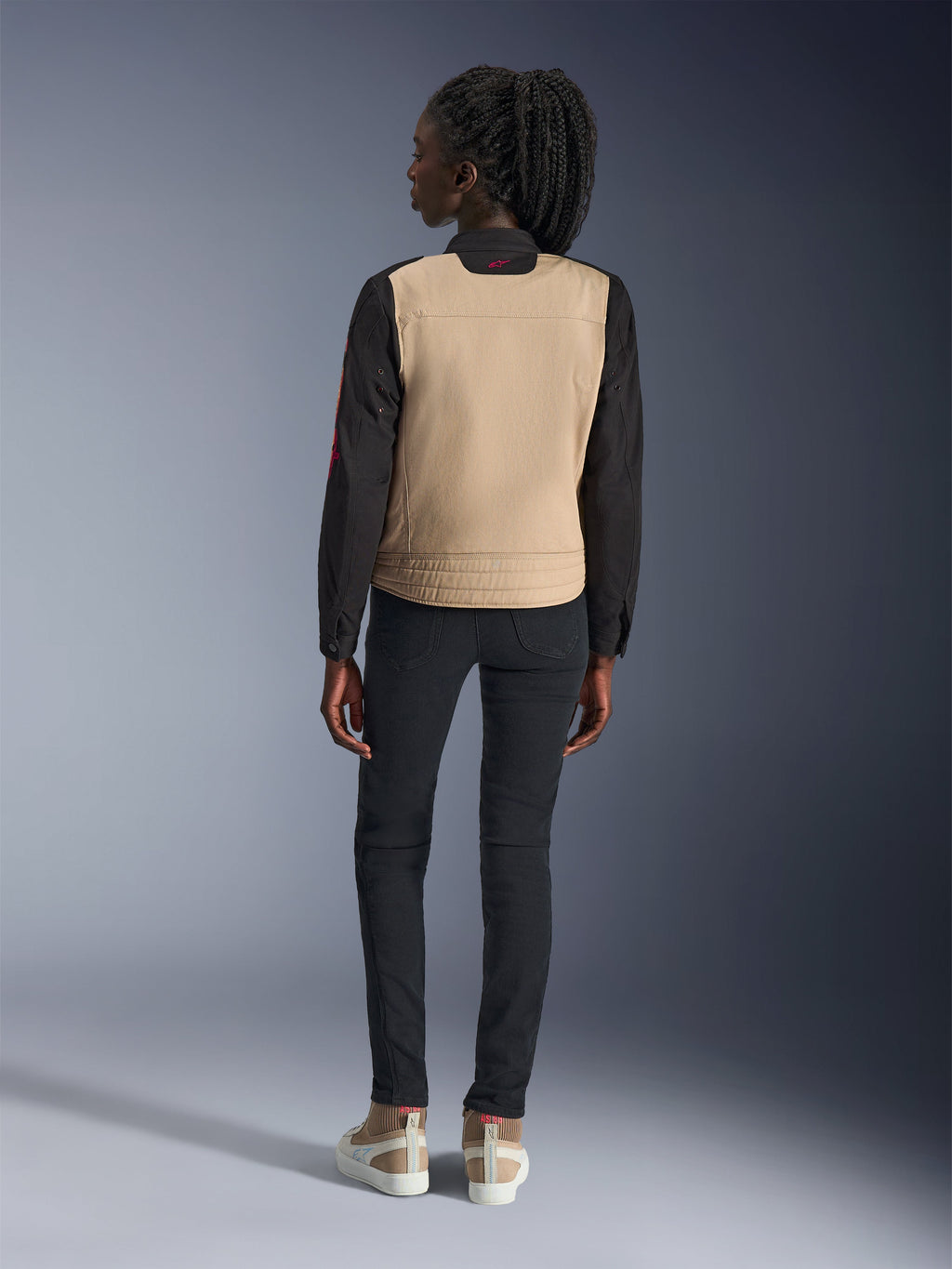 Kvinde iført Alpinestars Stella Grace jakke, Aluminum Black med tan torso og sorte ærmer, set bagfra, stylet med sorte slim-fit bukser og casual sneakers, urbant motorcykeludstyr til kvinder