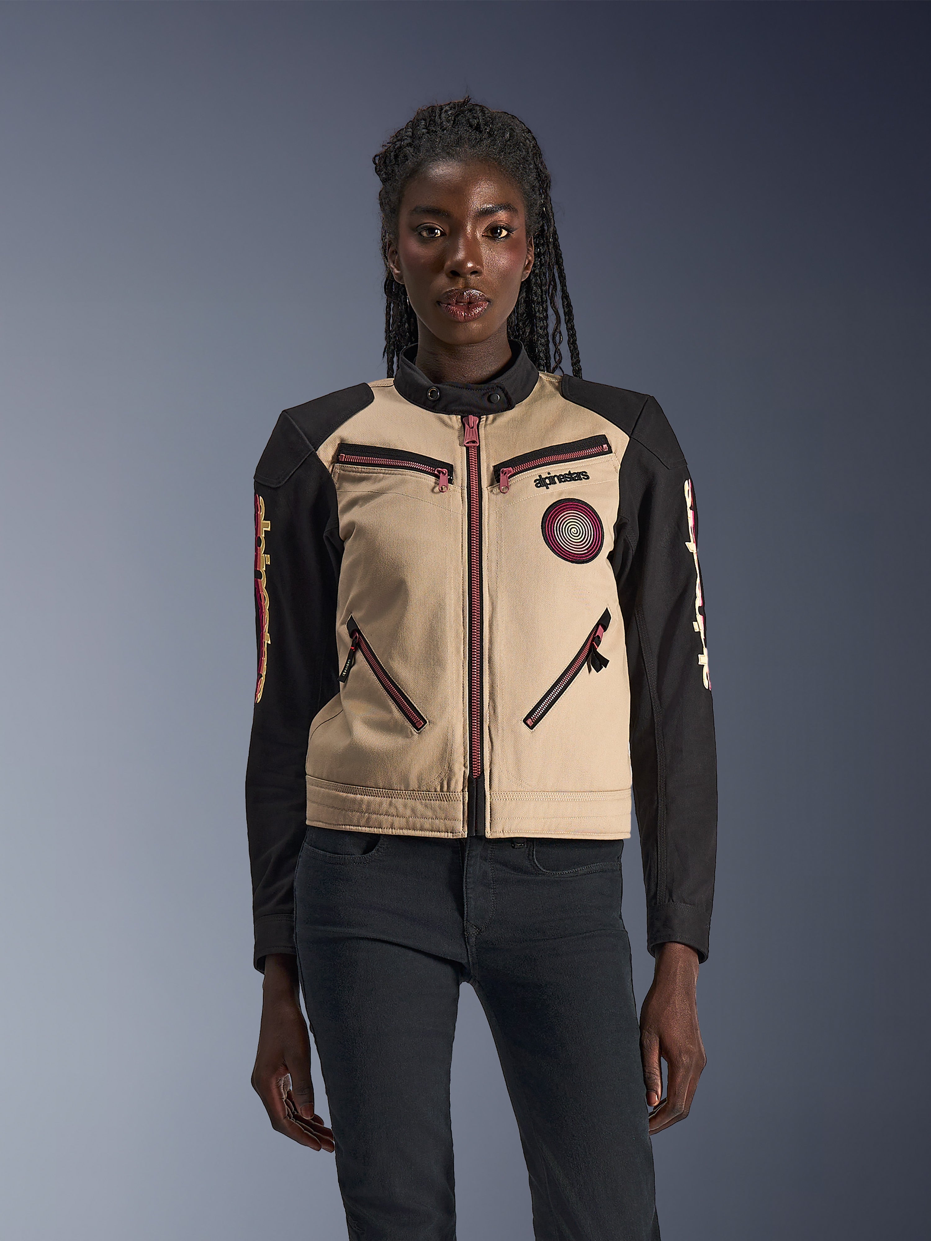 Alpinestars Stella Grace jakke, motorcykeljakke til damer, aluminum og sort, med et arbejdstøjsinspireret design, pink-røde lynlåsdetaljer, et spiralformet brystmærke og lange sorte ærmer med gul og rød grafik, båret af en model.