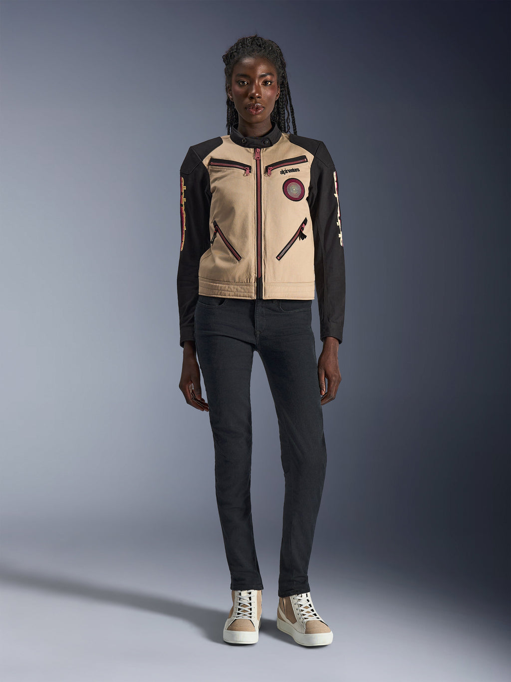 Alpinestars Stella Grace jakke til kvinder, urban motorcykeljakke, sandfarvet og sort tekstilkonstruktion, med lynlåse foran og broderet Oscar-spiralpatch, bæres med sorte bukser og high-top sneakers, moderne arbejdstøjsinspireret køreudstyr