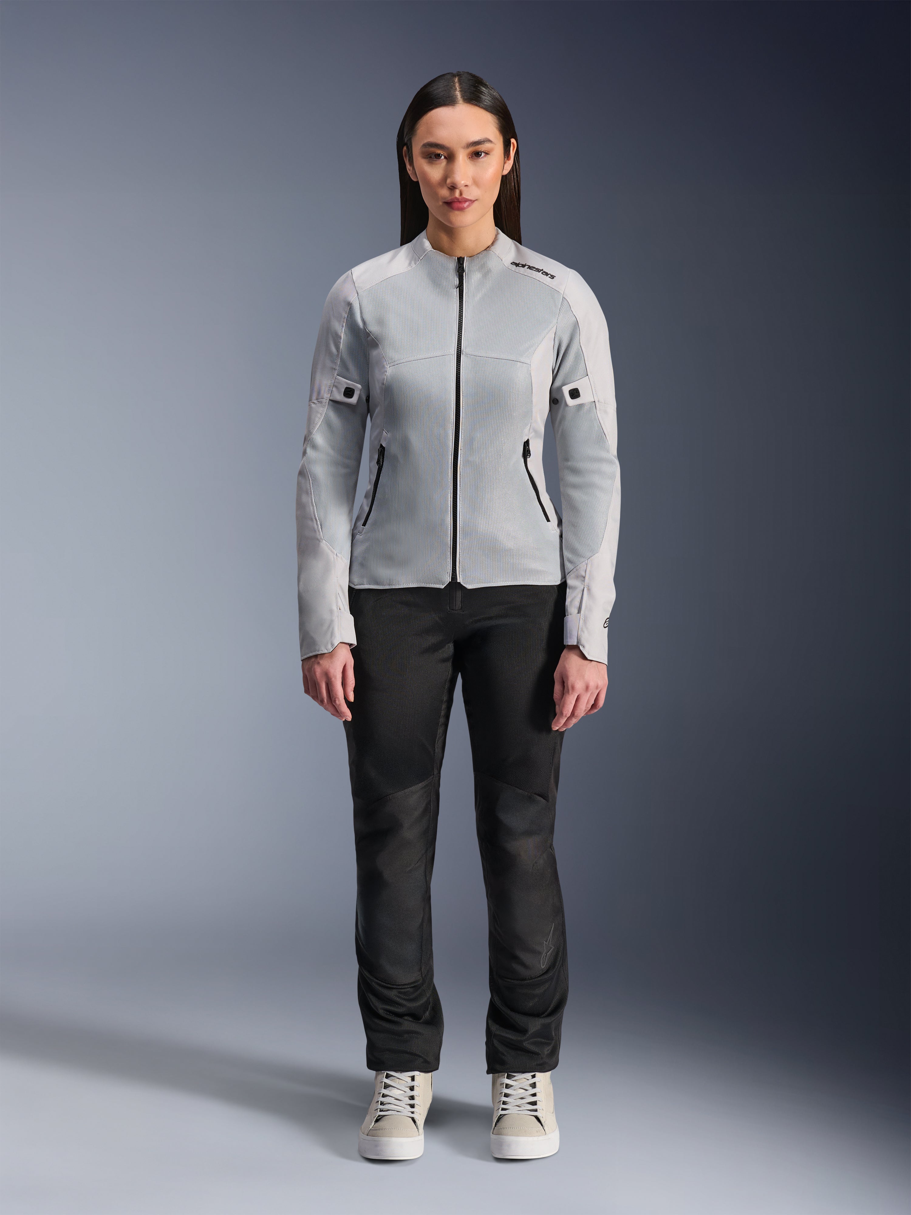 Kvinde iført Alpinestars Stella C-1 Air-jakke i sølv, set forfra, urban motorcykel tekstiljakke med tech mesh-paneler, stylet med sorte bukser og hvide sneakers, moderne køreudstyr