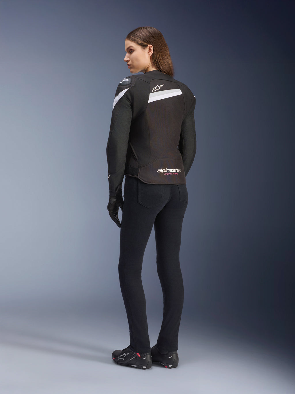 Alpinestars Stella T-GP Plus R V4 jakke, tekstiljakke til damer, sort og hvid, set bagfra på model, med hvide skulderdetaljer og Alpinestars-logo, båret med sorte bukser og kørehandsker, sport-fit design til vejkørsel