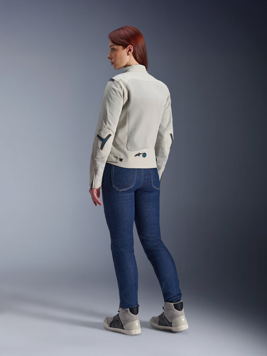 Kvinde iført Alpinestars Stella Circal-1 Air-jakke i Metal Gray og Methyl Blue, set bagfra som viser mesh-ventilationspaneler og neonblå accenter, kombineret med mørkeblå motorcykeljeans og grå sneakers, urbant motorcykel-livsstilsbillede.