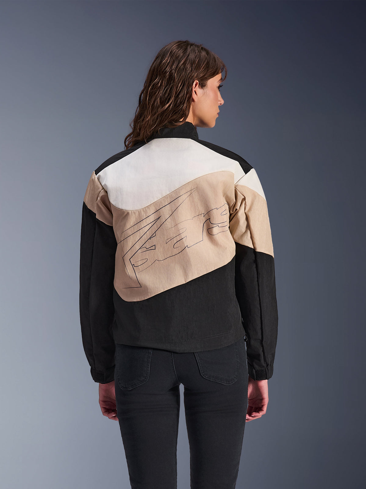 Alpinestars Stella Moshell-jakke, urban motorcykeljakke til kvinder, sort, sand og off-white, set bagfra på en model, asymmetrisk design med farveblokke og stor grafik på ryggen, moderne streetwear-inspireret beskyttelsesudstyr