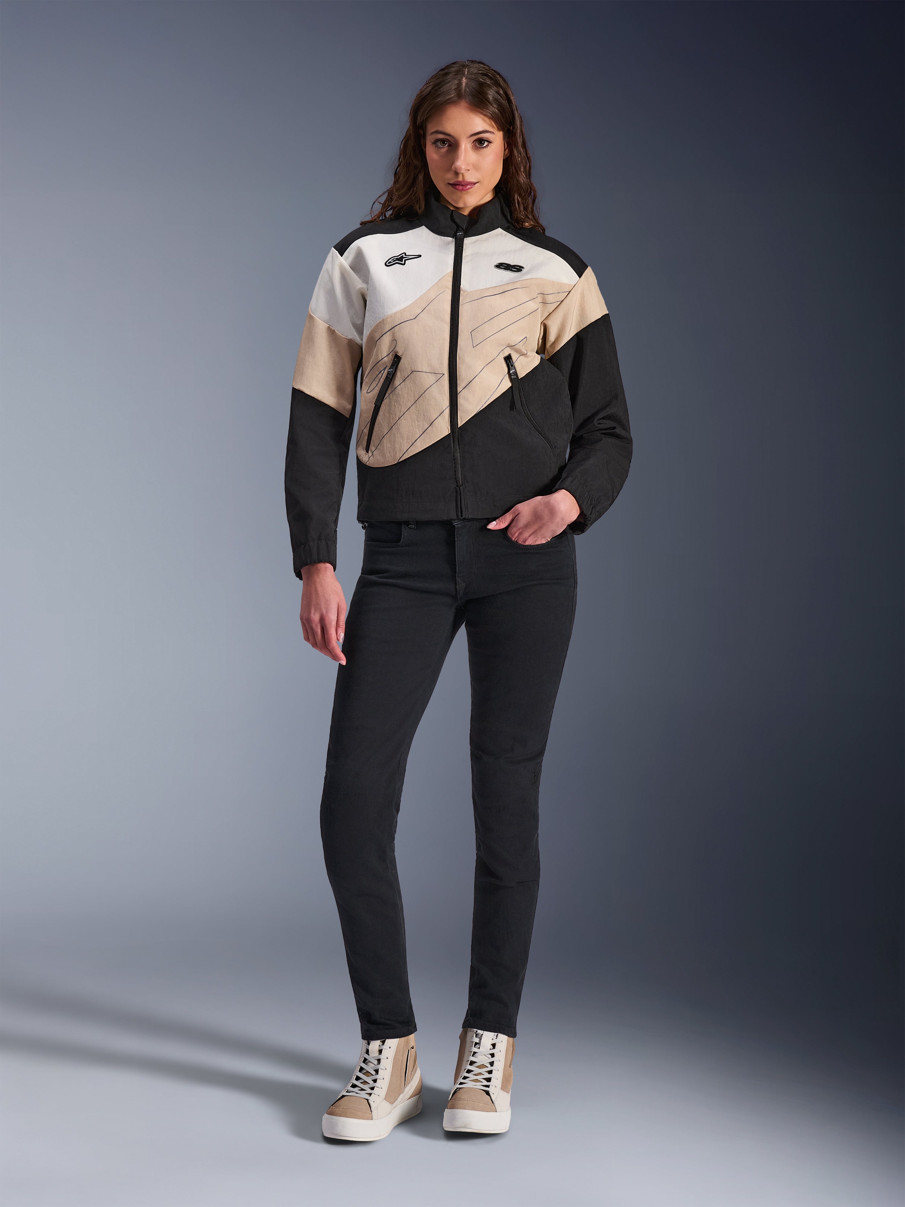 Alpinestars Stella Moshell-jakke til kvinder, urban motorcykeljakke med et asymmetrisk farveblokeret design i sort, sand og off-white, båret med sorte slim-fit bukser og sandfarvede high-top sneakers, moderne streetwear-æstetik til kørere