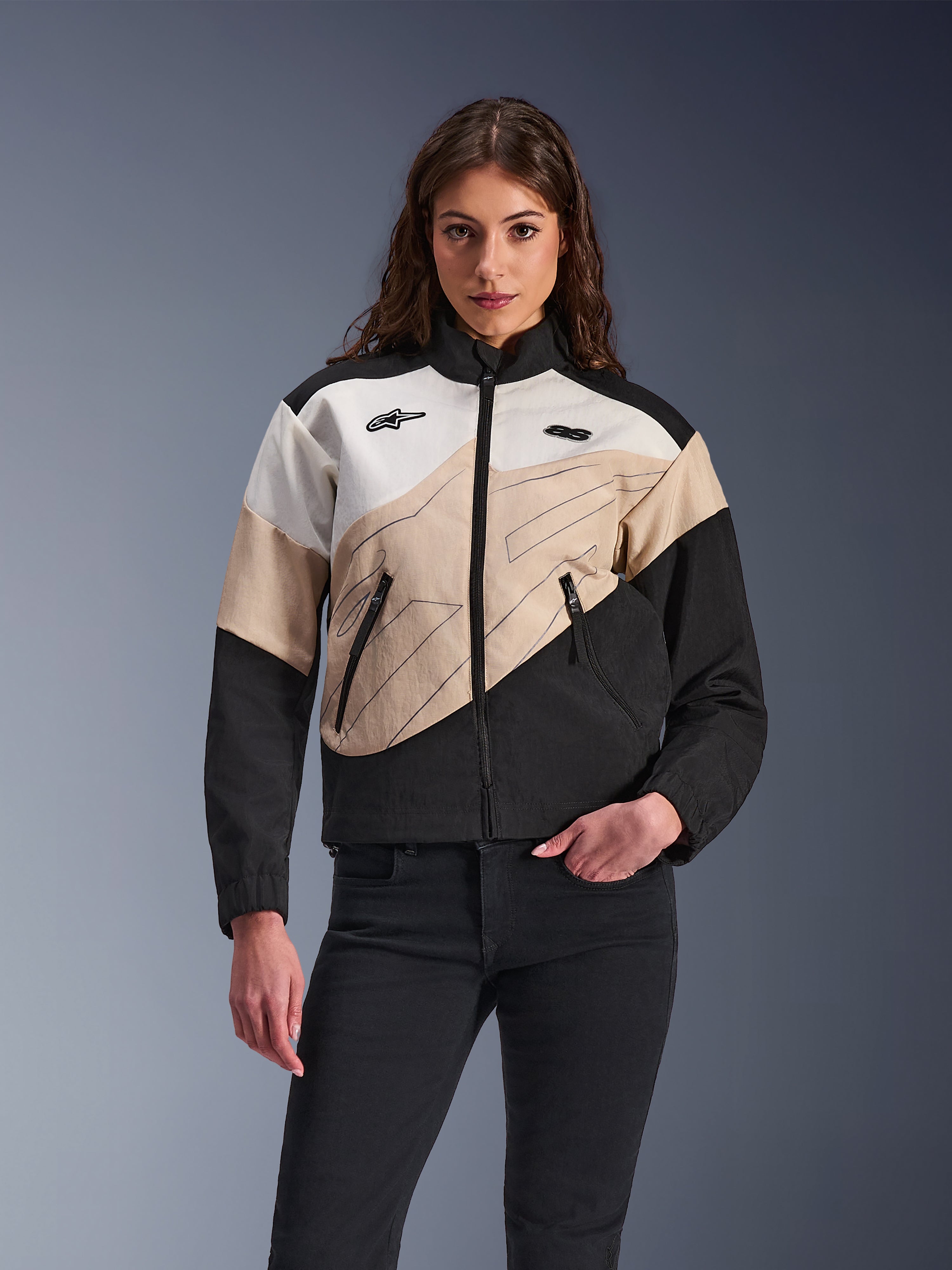 Alpinestars Stella Moshell jakke, urban motorcykeljakke til kvinder, asymmetrisk color-blocked design i sort, sand og off-white, med lynlås foran og sidelommer med lynlås, vist med sorte bukser for et moderne streetwear-look.