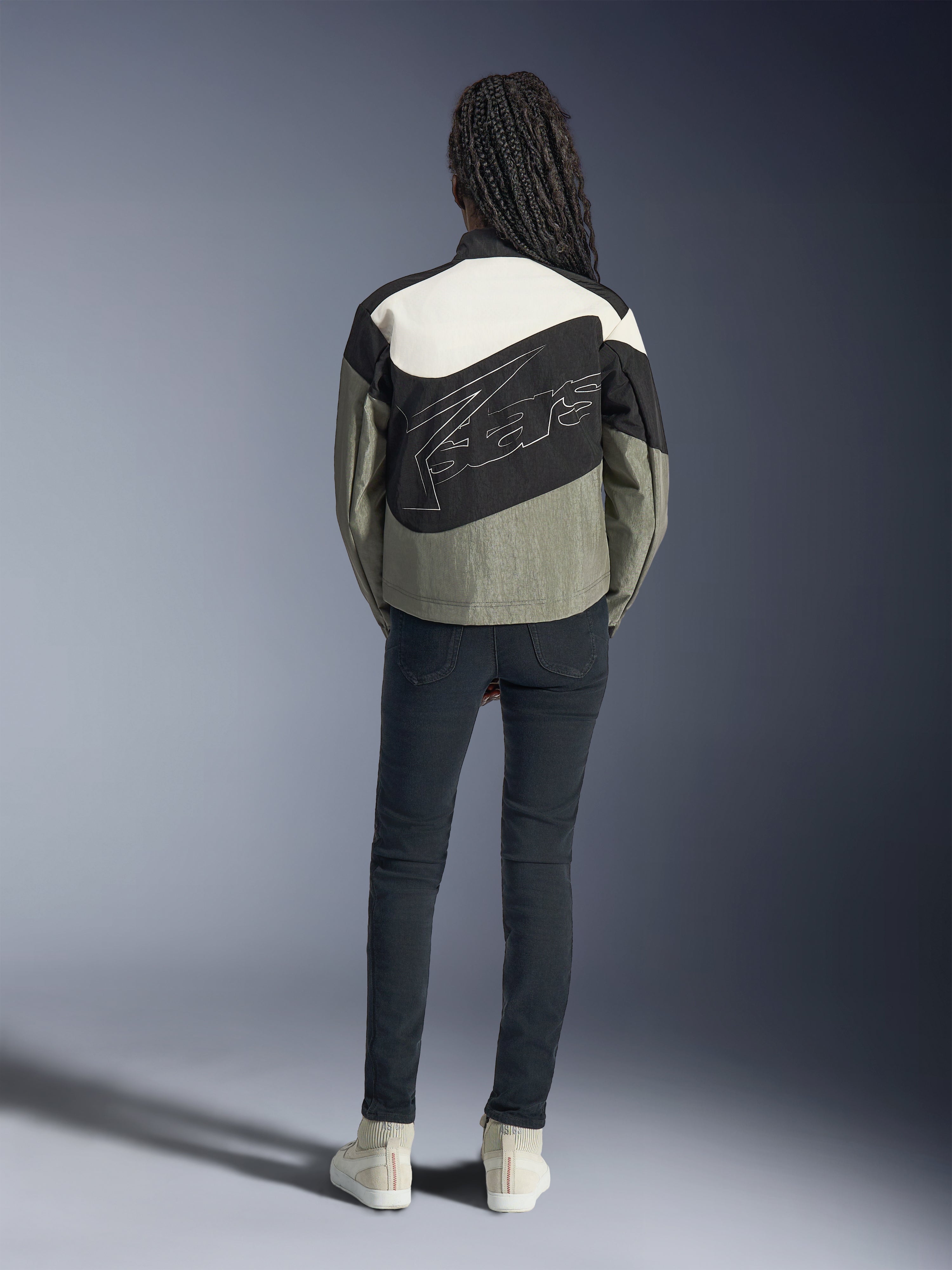 Model set bagfra iført Alpinestars Stella Moshell-jakke i sort, grå og off-white, med et asymmetrisk design og stort stiliseret konturlogo på ryggen, kombineret med mørke slim-fit jeans og hvide sneakers, urbant streetwear-inspireret motorcykeltøj til kvinder.